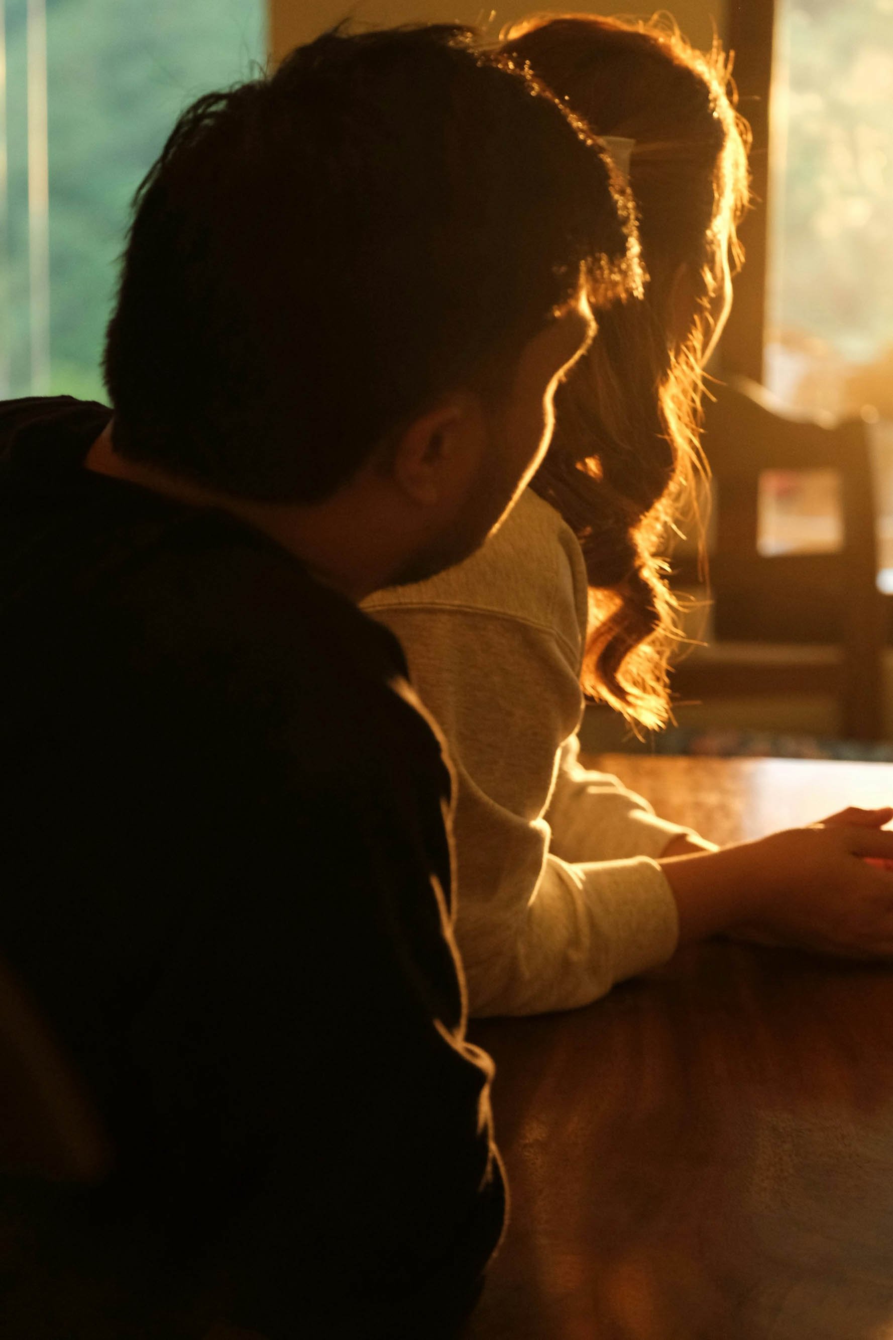 Two people sitting side by side at a table, with warm sunlight illuminating their hair from behind, inside a room with a window in the background.