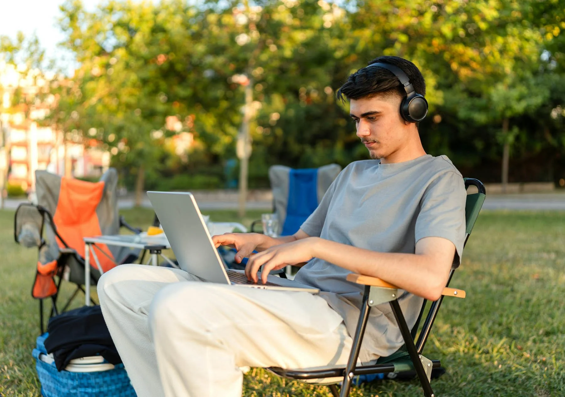 A young man wearing a gray t-shirt and white pants sitting outdoors on a folding chair, working on a laptop, with headphones on, in a park with trees and greenery in the background.