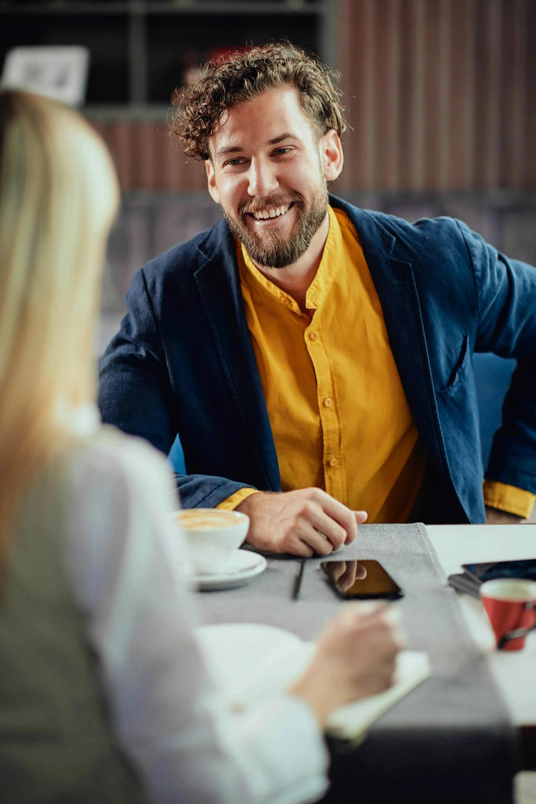 A smiling man with curly hair and a beard, wearing a yellow shirt and a denim jacket, engaged in conversation.