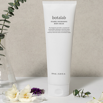 A white tube of Botalab Suamel Nourishing Body Cream standing upright on a white surface, with flowers and greenery around it, against a neutral background.