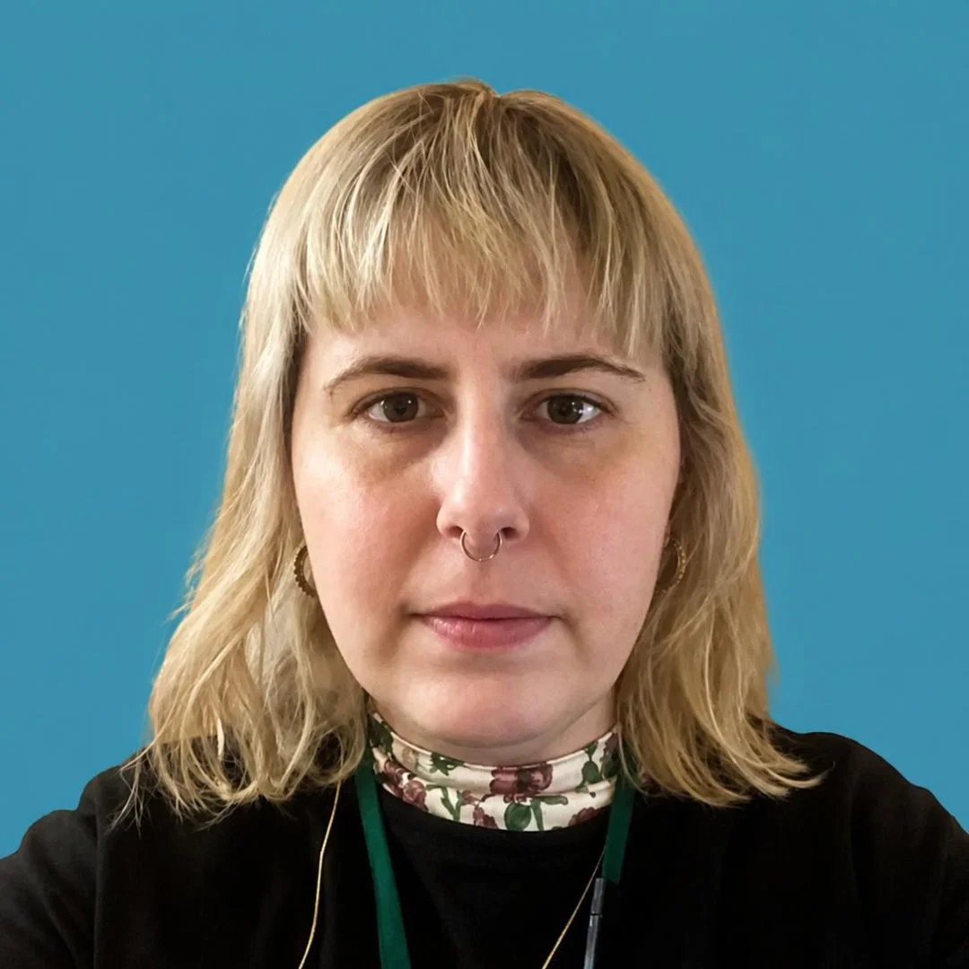 A woman with shoulder-length blonde hair, wearing a black top, floral turtleneck, and earphones, standing against a blue background.
