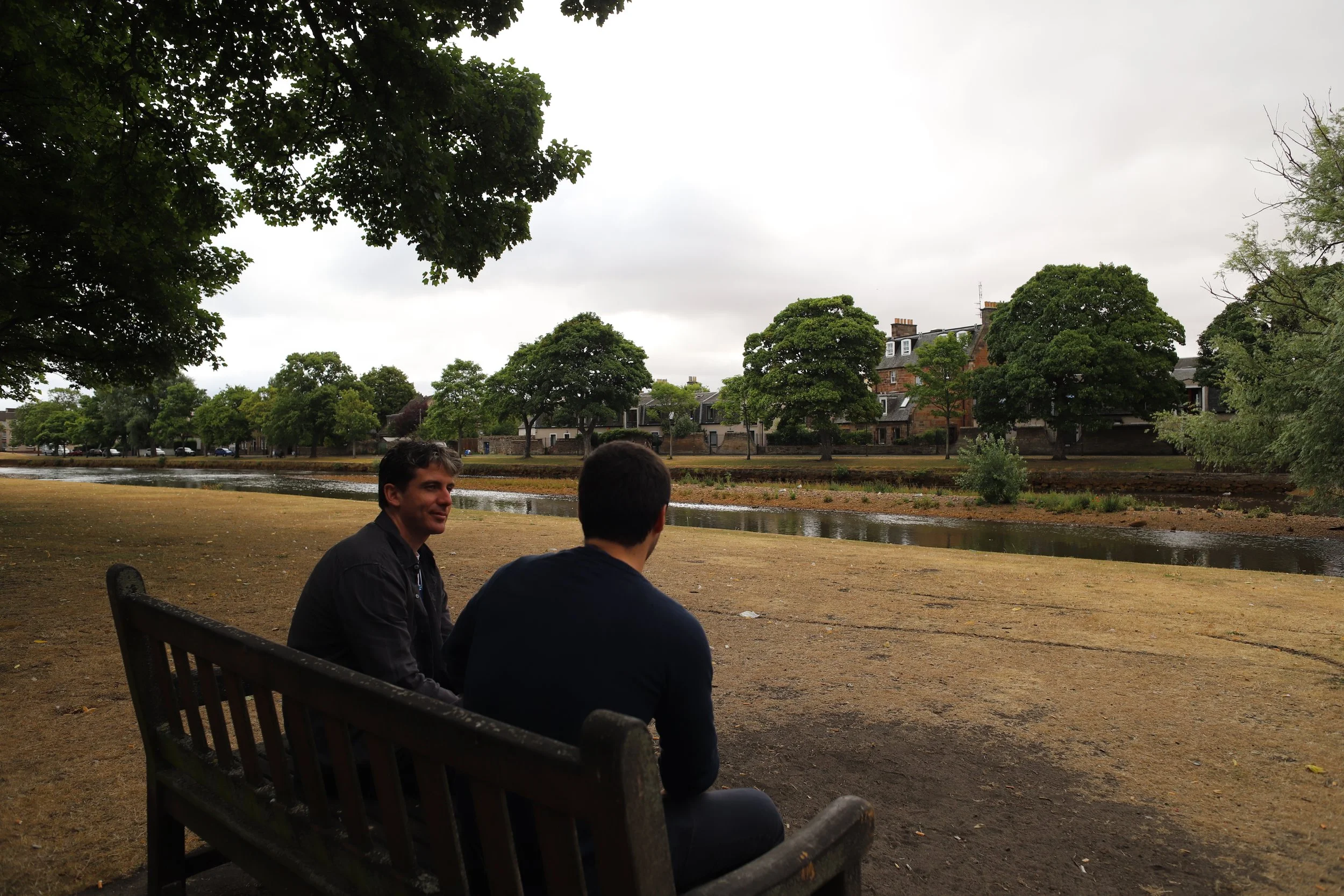 Two men sitting on a bench by a river, with large leafy trees and buildings in the background under a cloudy sky.