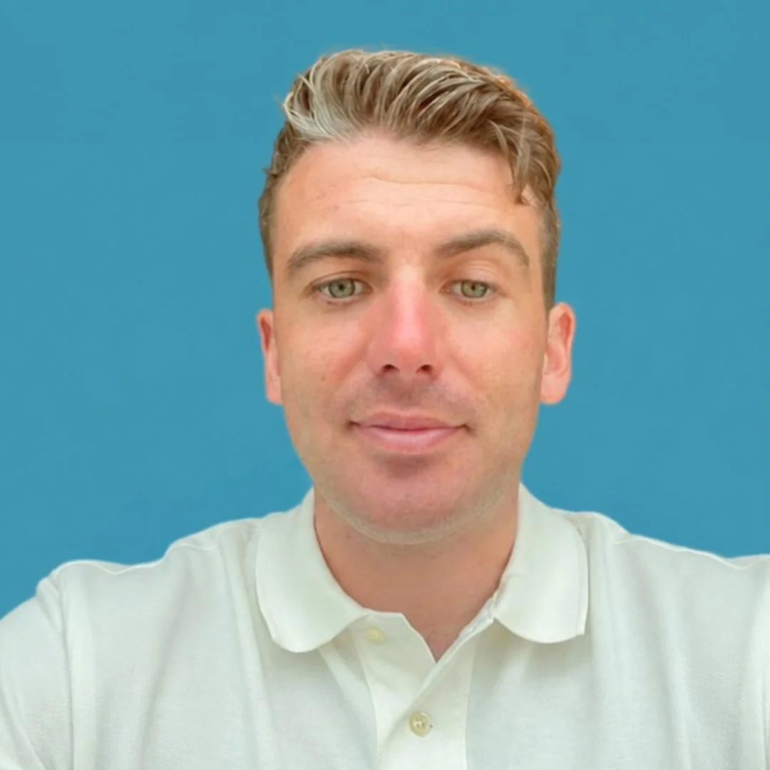 A man with light hair and fair skin is taking a selfie against a blue background, wearing a white collared shirt.