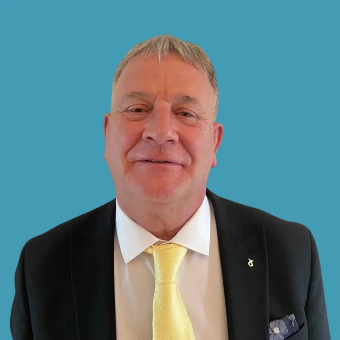 Portrait of a middle-aged man smiling, dressed in a black suit with a yellow tie, against a solid blue background.