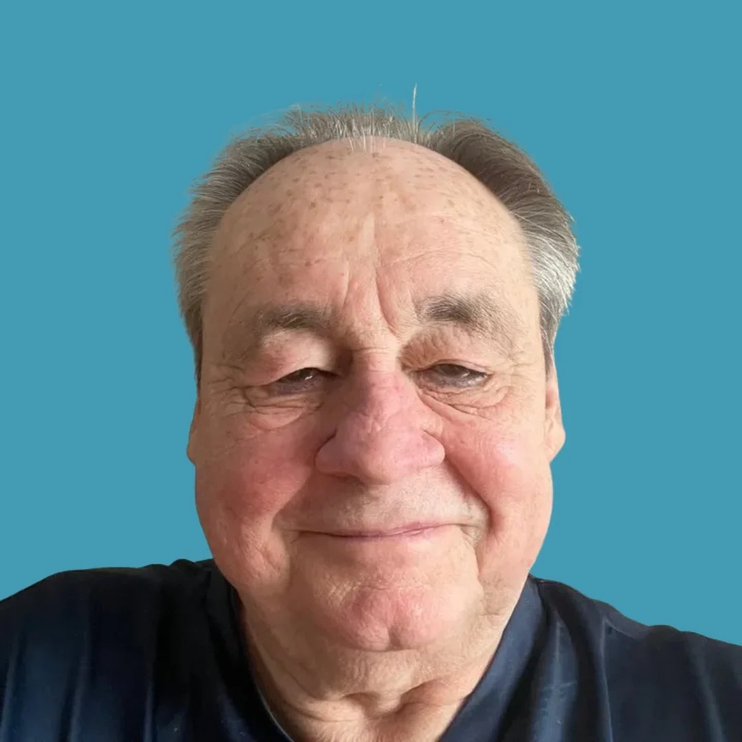 Close-up of an elderly man with short, gray hair and a warm smile, wearing a dark shirt, against a solid blue background.