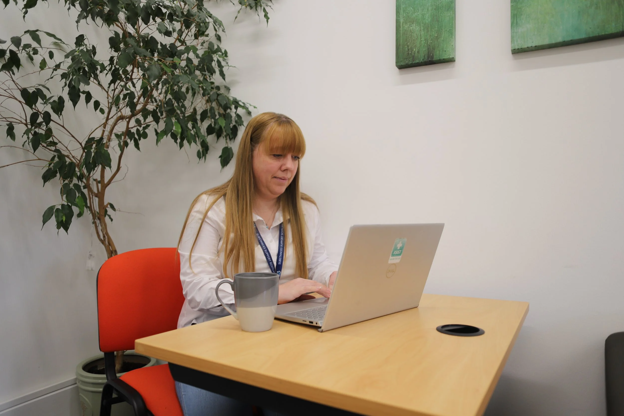 Woman with long blonde hair working on a laptop at a wooden table in an office with white walls, green wall art, potted plant, and orange and black chairs.