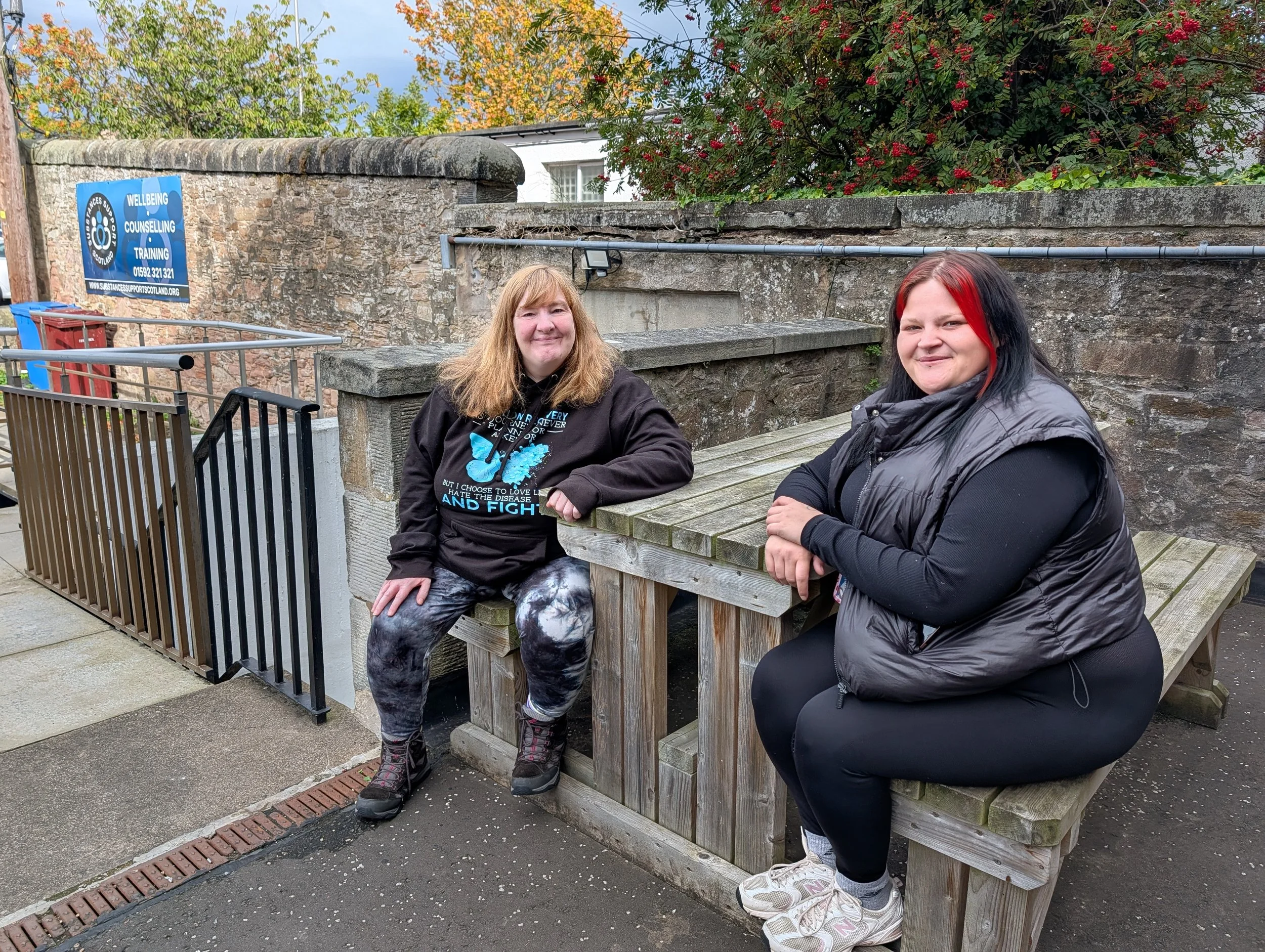 Two smiling women sitting on a wooden bench outdoors, with a stone wall behind them and trees with red and green leaves overhead.
