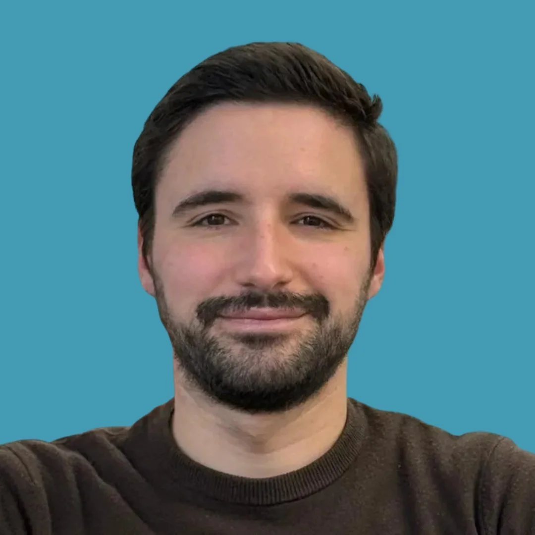 Portrait of a man with dark hair and a beard, smiling, against a solid blue background.