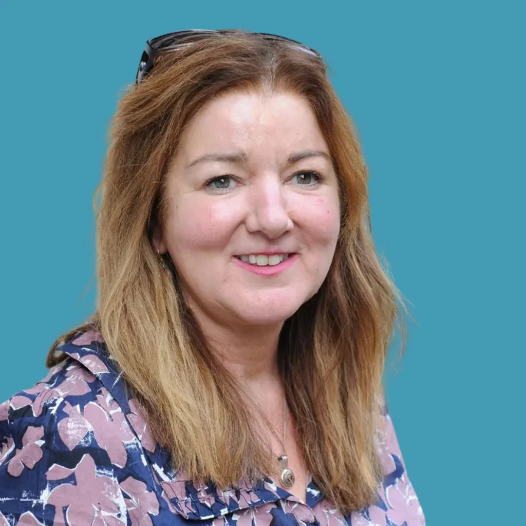 Portrait of a woman with shoulder-length reddish-brown hair, wearing a blue patterned blouse and sunglasses on her head, against a solid teal background.