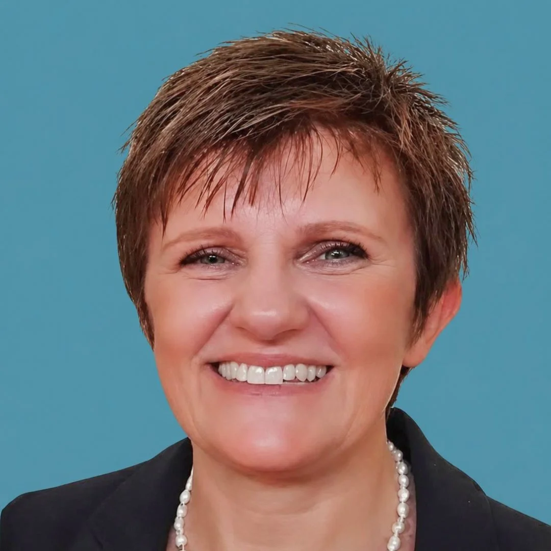 A woman with short, brown hair, smiling, wearing a pearl necklace and a black blazer against a blue background.