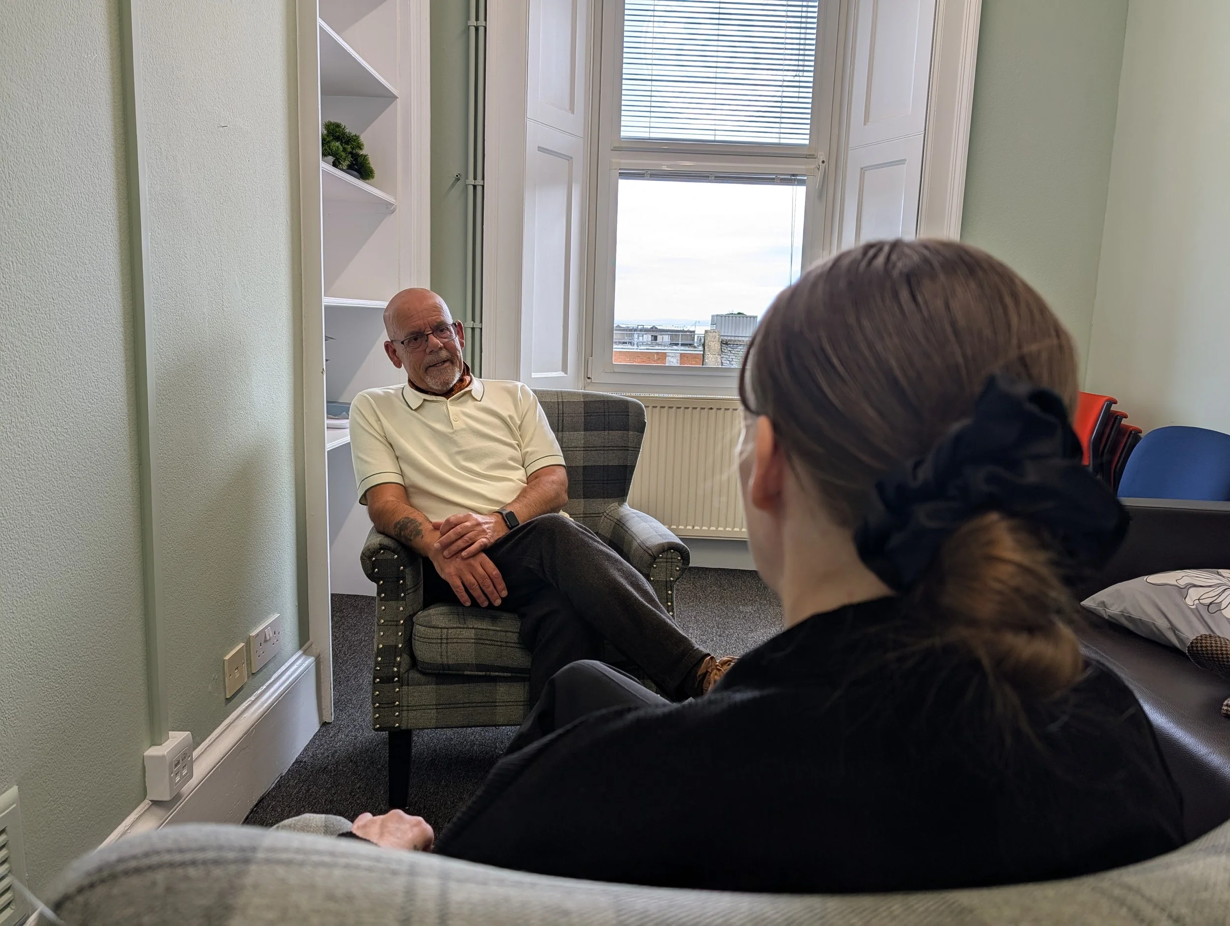 Two men are sitting and talking in a room with green walls and a window with blinds. One man has a beard and glasses, wearing a light-colored polo shirt, while the other has short hair and is wearing a dark hoodie. There are chairs stacked against the wall on the right side of the image.