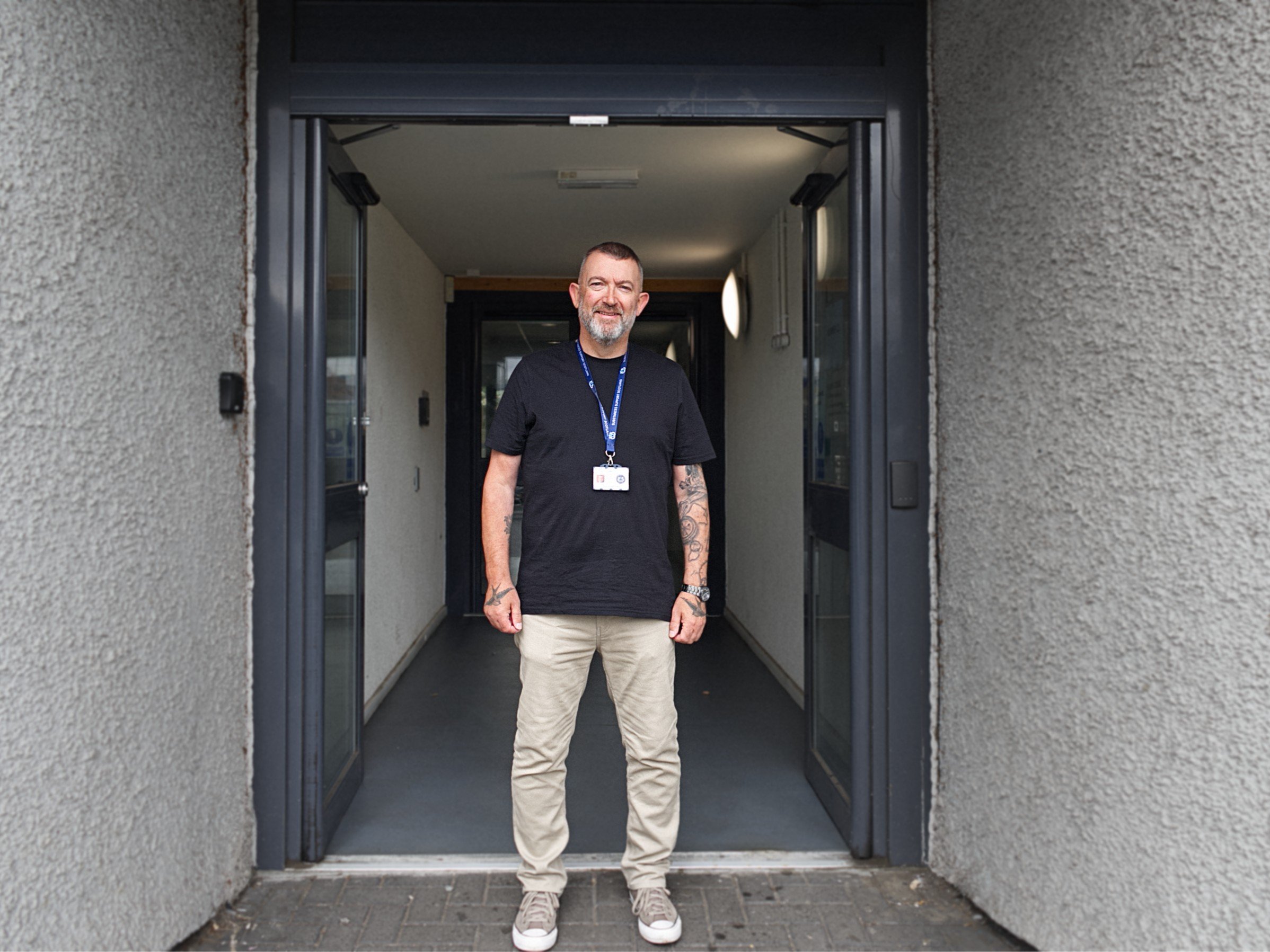 A man with short hair, a beard, and tattoos on his arms, standing at the entrance of a building with a black shirt, beige pants, and sneakers, smiling at the camera.