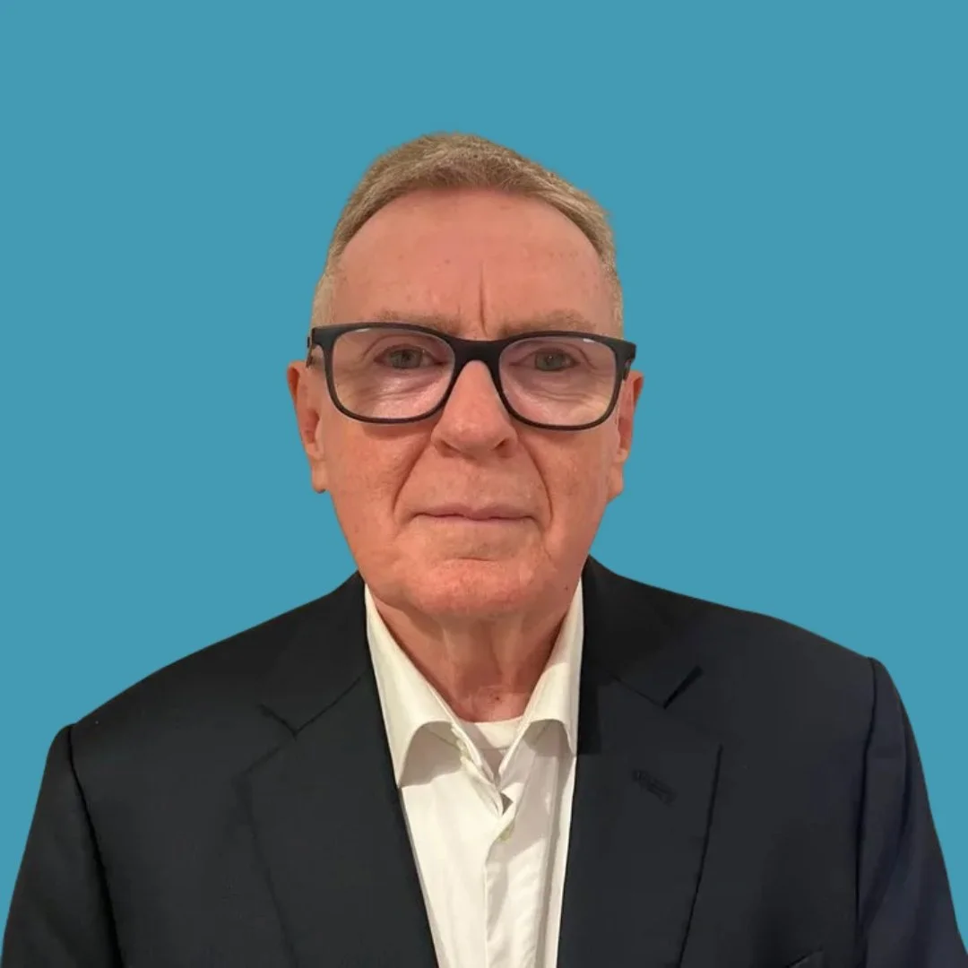 A man in a black suit, white shirt, and black glasses stands against a blue background.