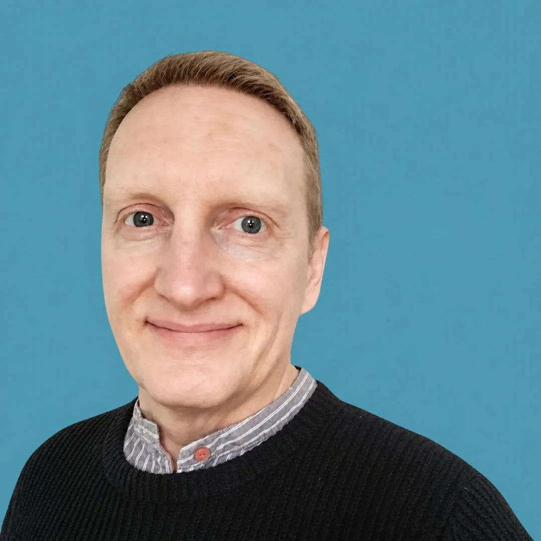 Close-up portrait of a smiling man with short red hair, wearing a black sweater over a striped collared shirt, against a blue background.