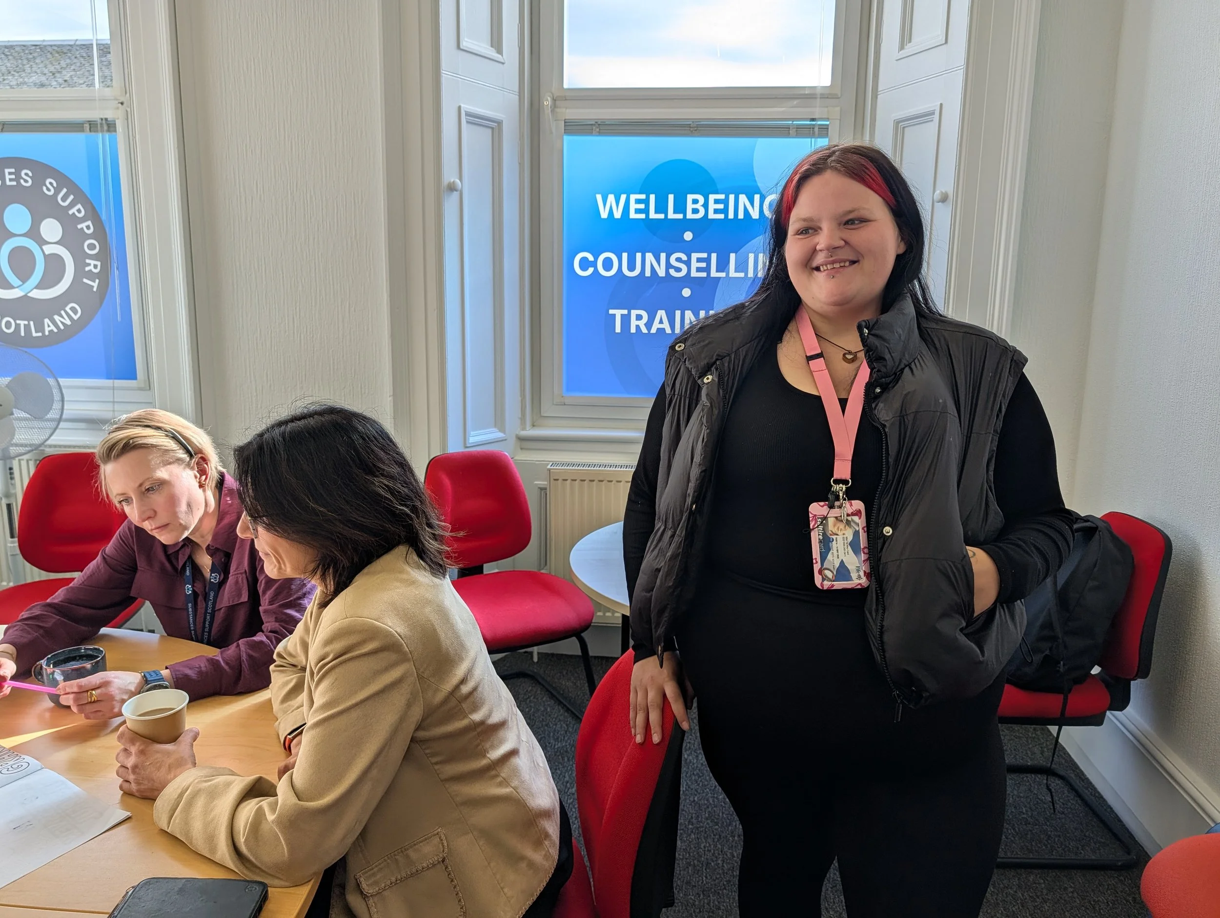 Group of people at a meeting table with a woman standing, smiling, wearing a black shirt and black vest, with a lanyard around her neck. Windows in the background display signs about wellbeing, support, counseling, and training.