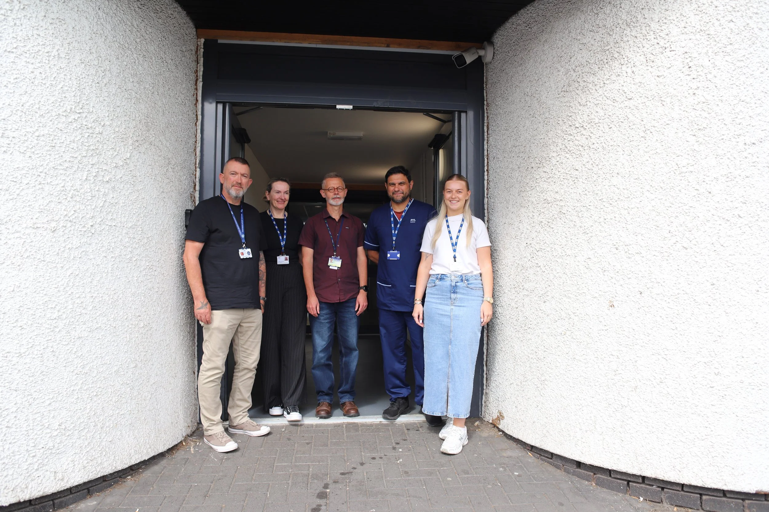 Group of five diverse people standing together in front of a building entrance, smiling, with a security camera visible on the wall.