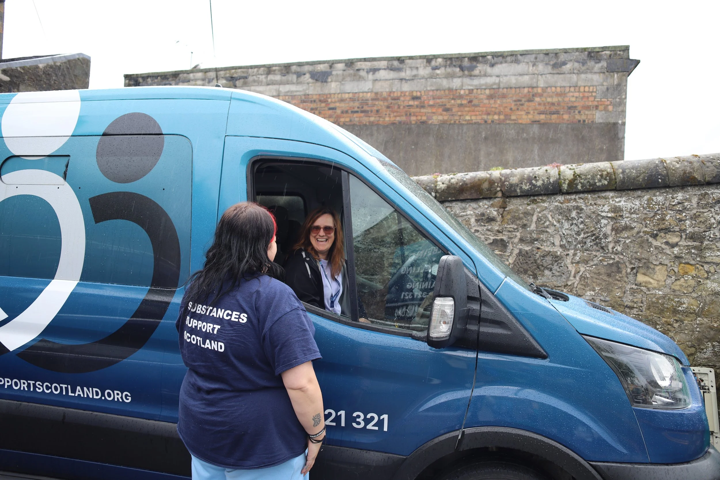 A woman with long hair, wearing sunglasses, is sitting inside a blue support vehicle with the words 'SUBSTANCES SUPPORT SCOTLAND' on the side. Another woman with dark hair, wearing a navy blue shirt with similar support text, is standing outside and talking to her. The vehicle is parked in front of a stone wall and part of a building, with a cloudy sky overhead.