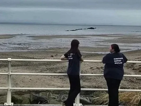 Two women sitting on a bench by the beach, wearing matching navy blue shirts with white text, facing the ocean under a cloudy sky.