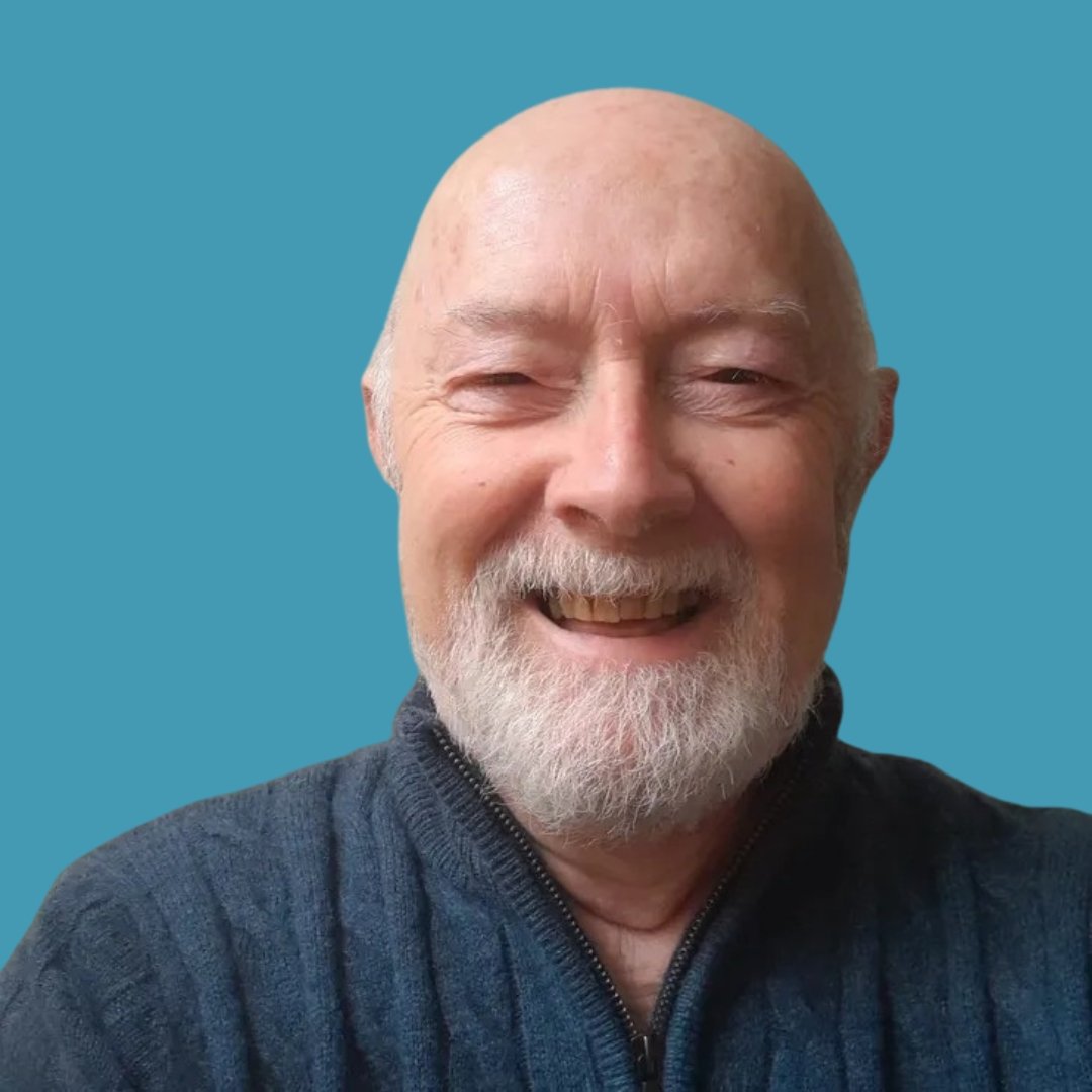An elderly man with a bald head and white beard, smiling, wearing a dark sweater, against a blue background.
