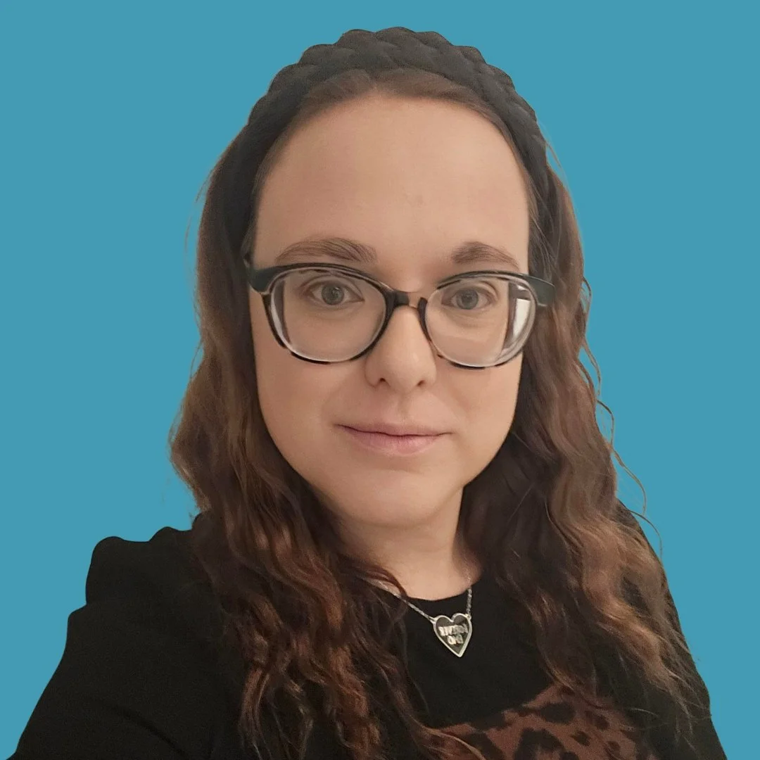 A woman with long wavy brown hair, glasses, and a black top with a leopard print section, standing against a blue background.