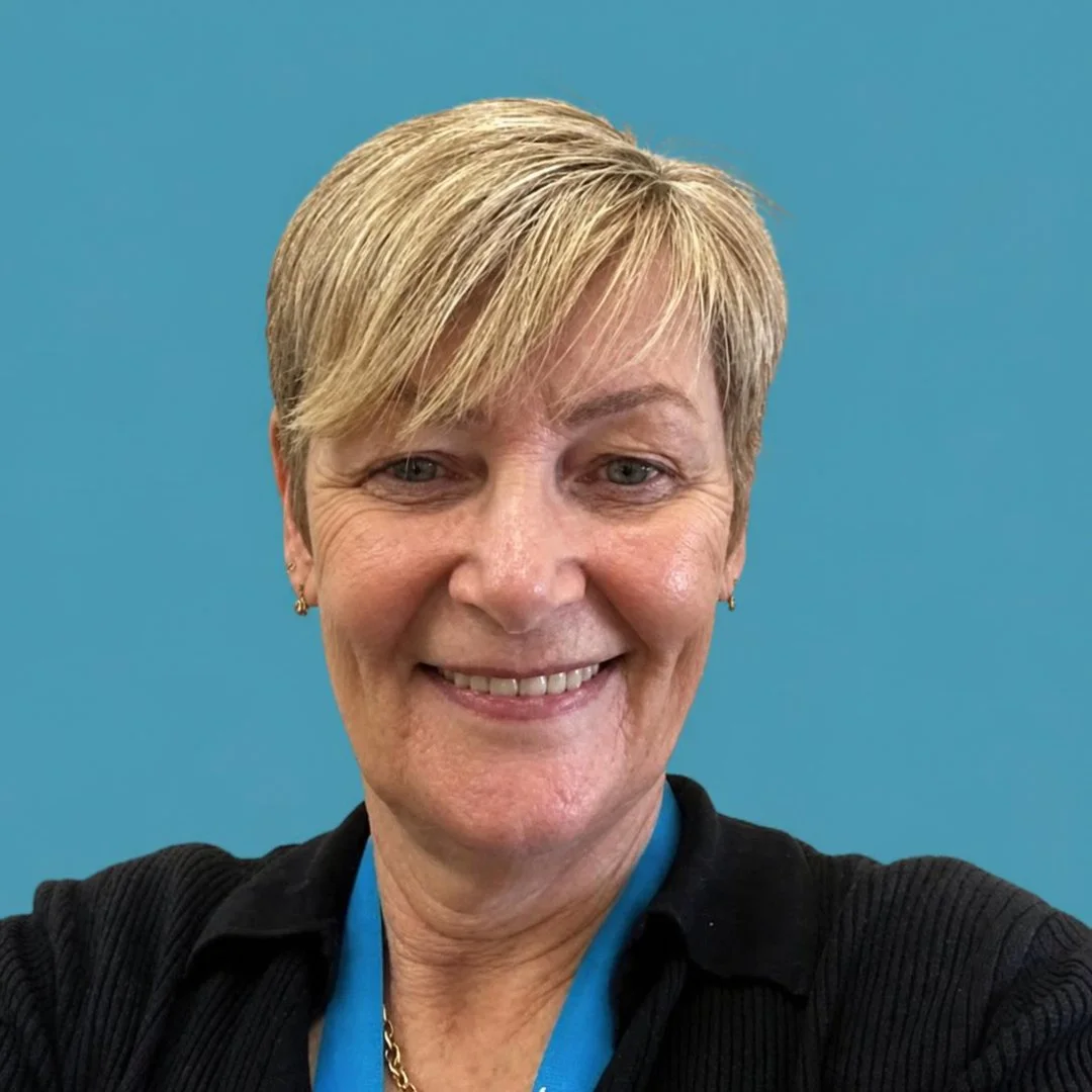 A smiling woman with short blonde hair, wearing a black top and a blue lanyard, in front of a plain blue background.