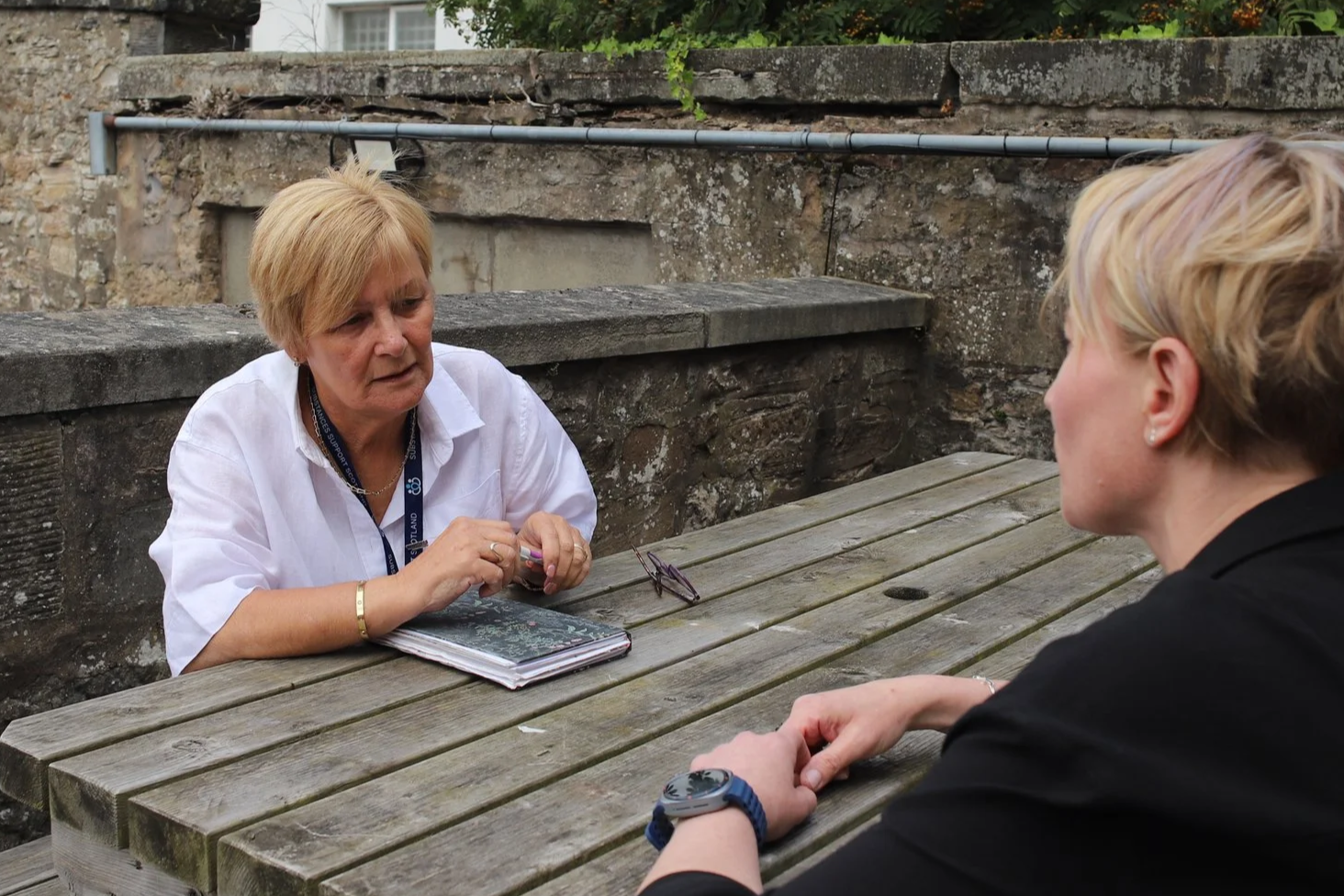 Two women sitting at a weathered wooden outdoor table, engaged in a serious conversation. One woman has short blonde hair, wears a white shirt, and has a notebook and glasses on the table. The other woman, also with short blonde hair, wears a black top and a watch.