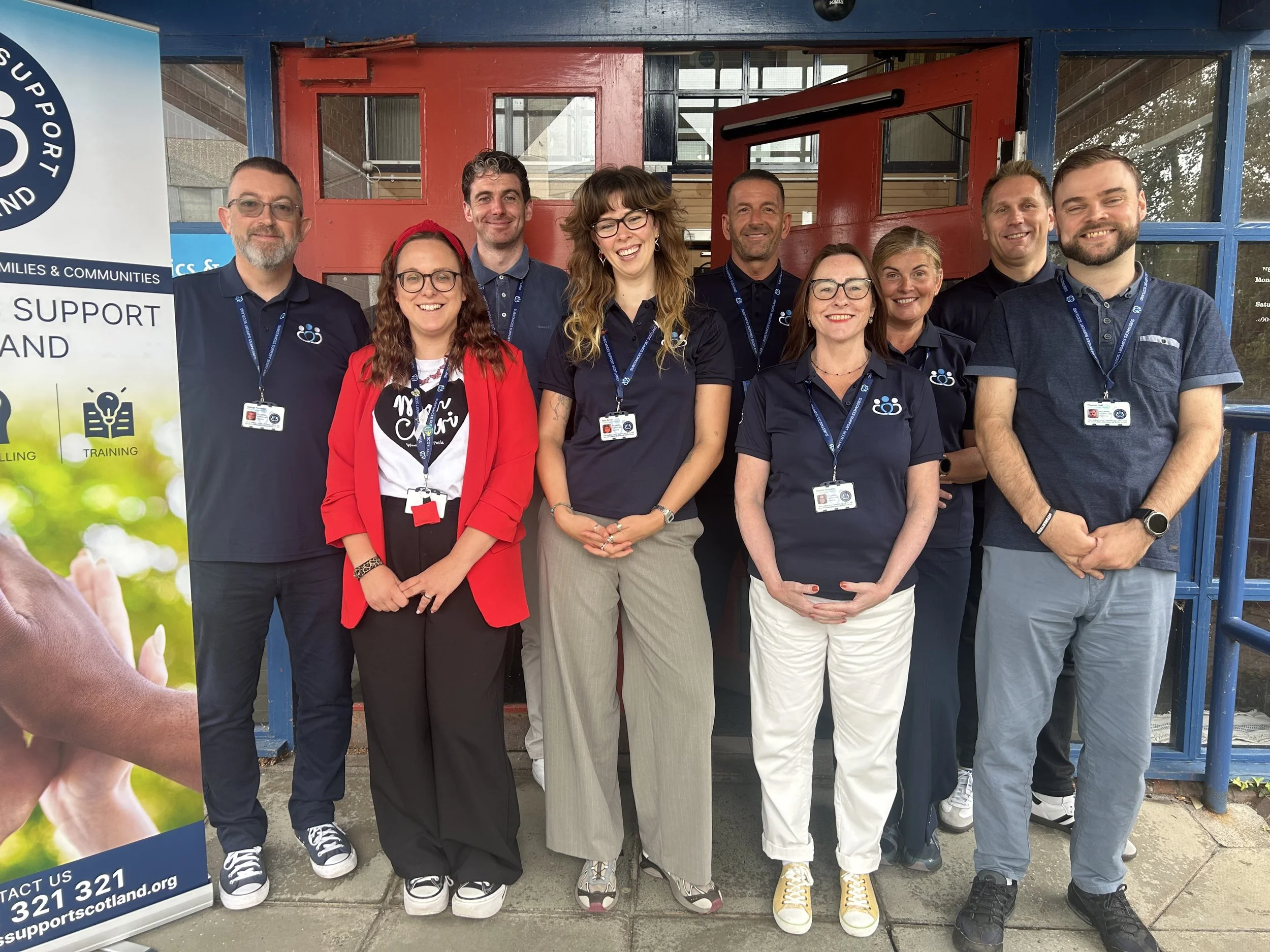A group of nine people standing outside a building, smiling and wearing navy blue shirts with an organization logo, with a support organization banner on the side.