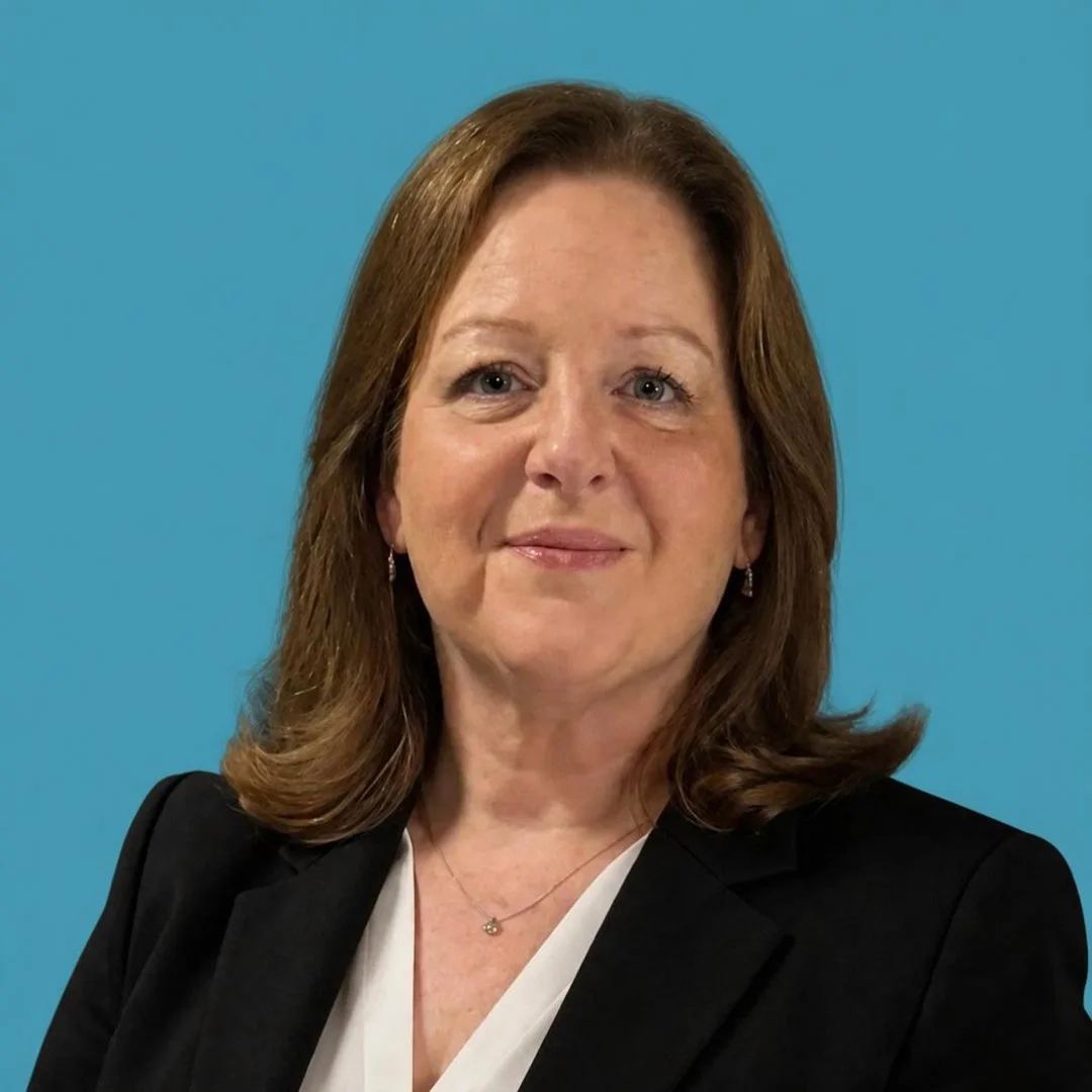 A professional woman with shoulder-length brown hair, wearing a black blazer over a white top, standing against a blue background.