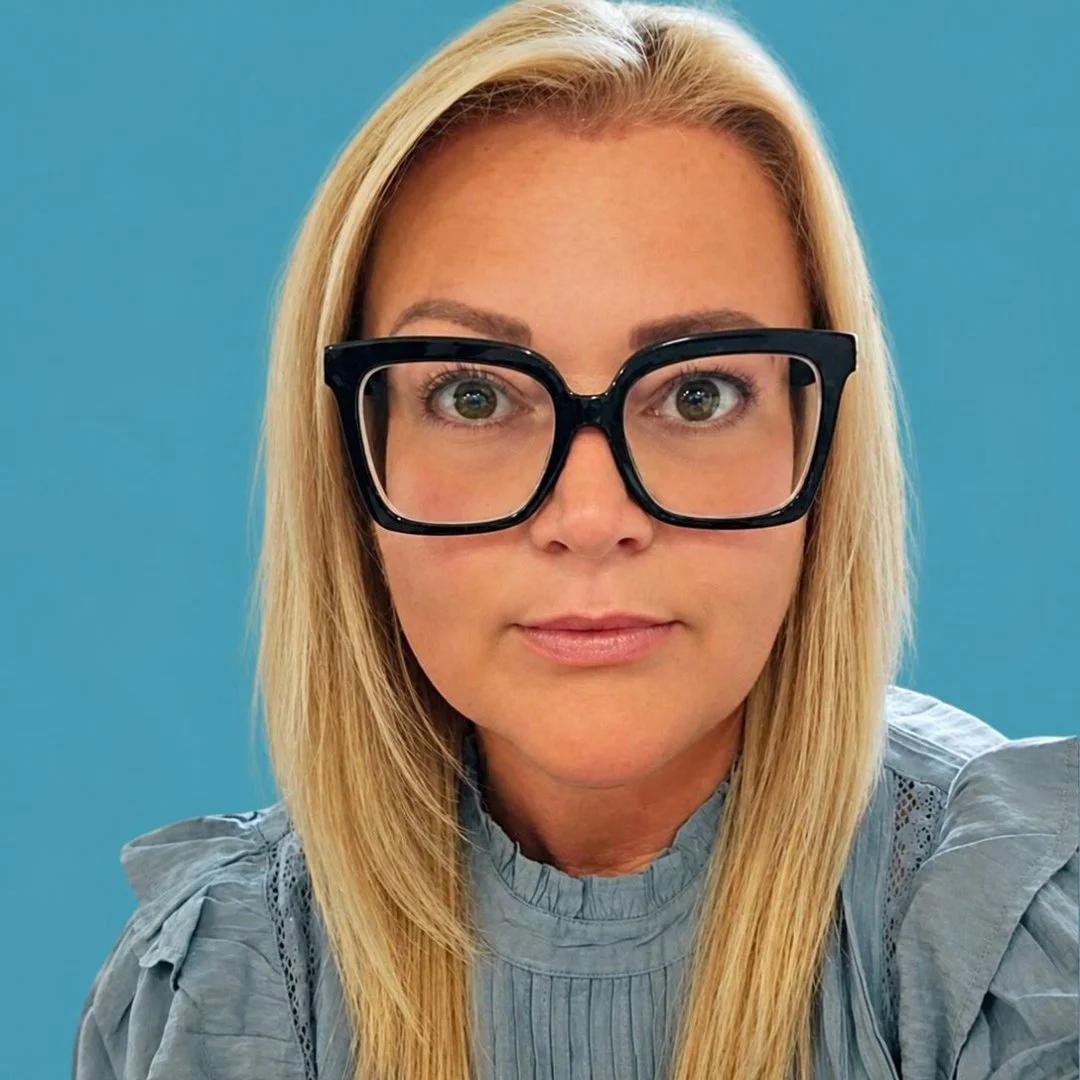 Close-up of a woman with blonde hair wearing large black glasses and a gray top, set against a blue background.