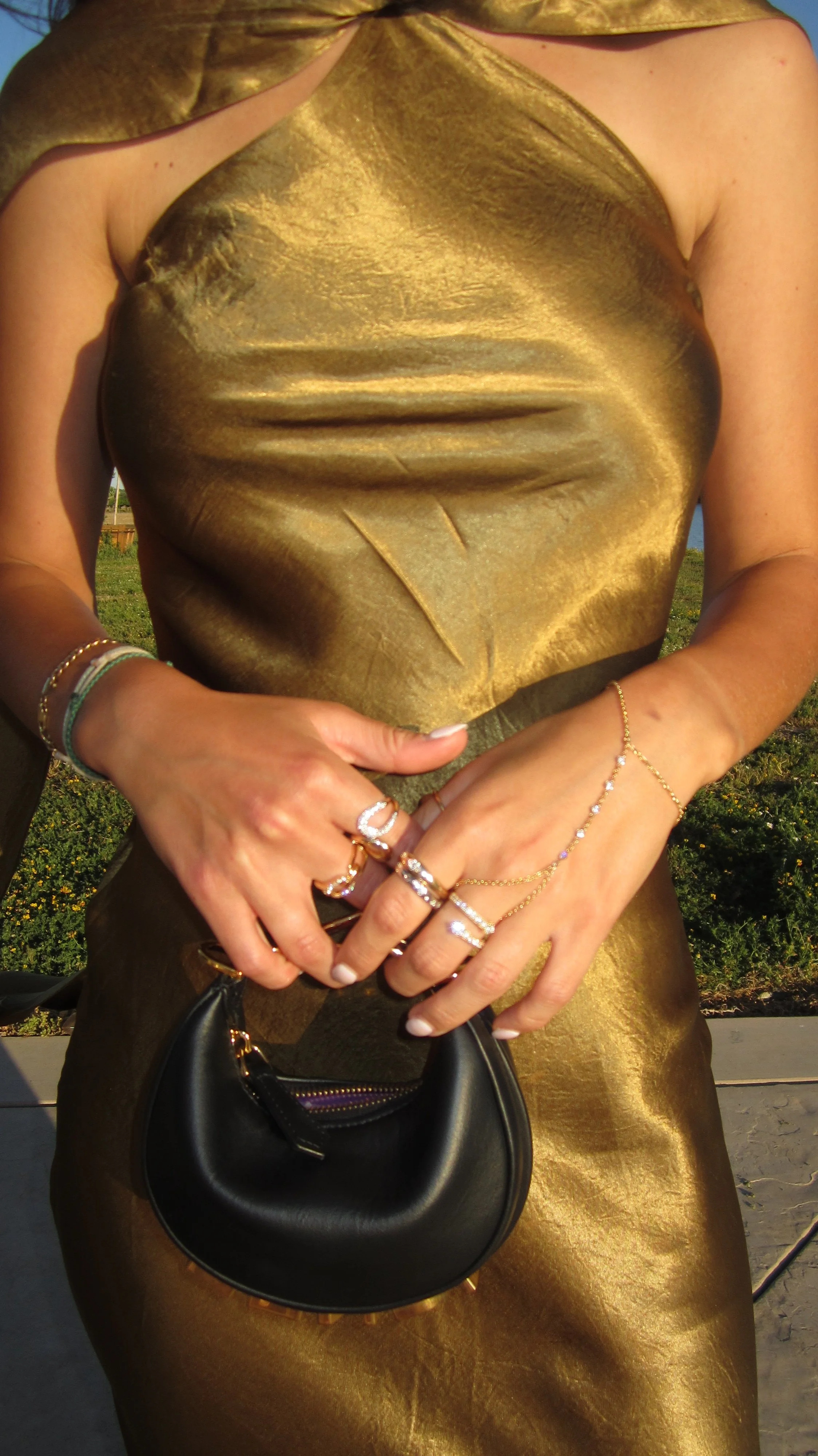 Person wearing a shiny gold dress, holding a small black handbag. Multiple rings and bracelets on their hands, standing outdoors with a background of grass and blue sky.