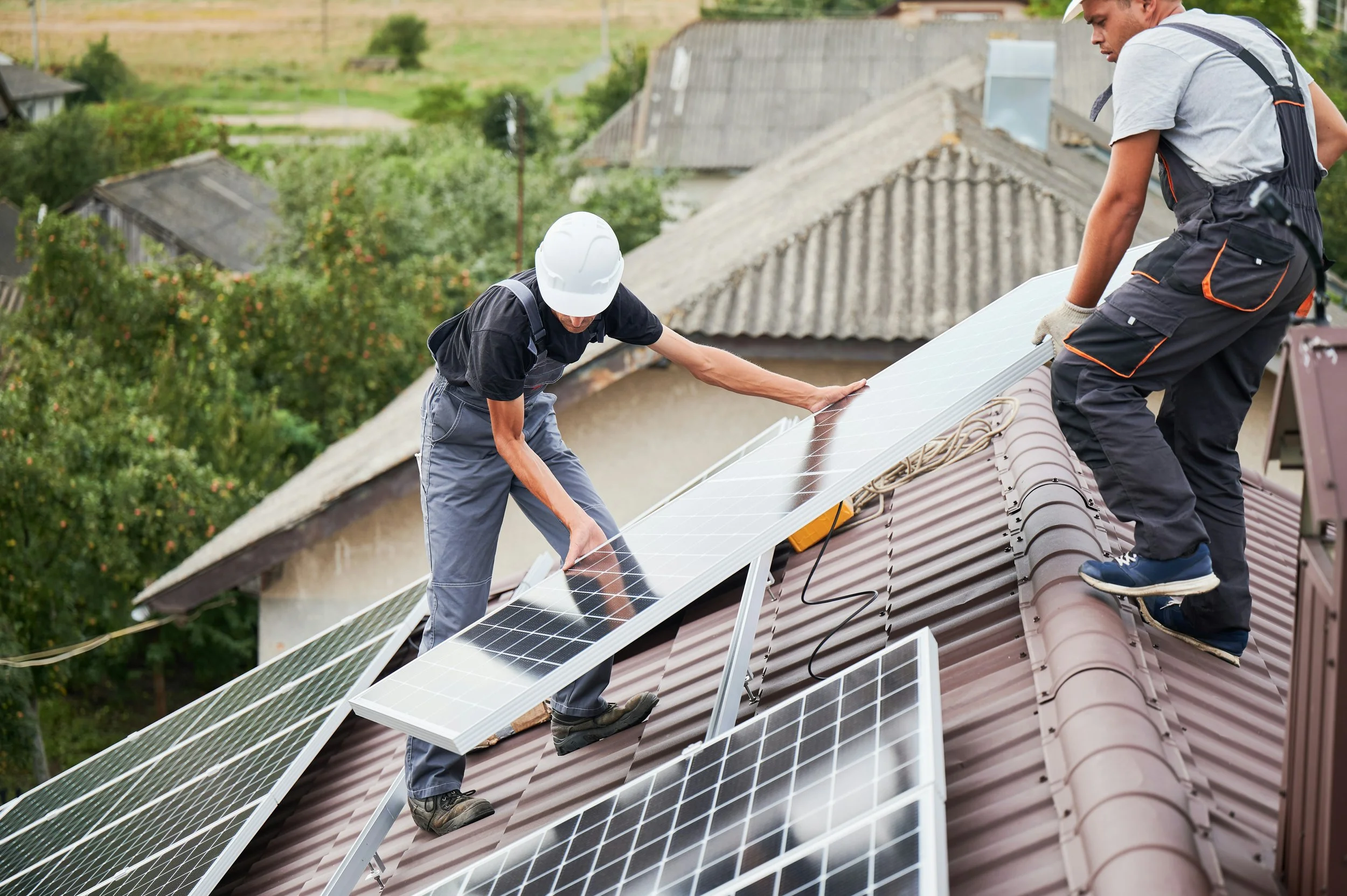 Two workers installing solar panels on a sloped roof of a residential house. One is wearing a white helmet and both are using safety harnesses.