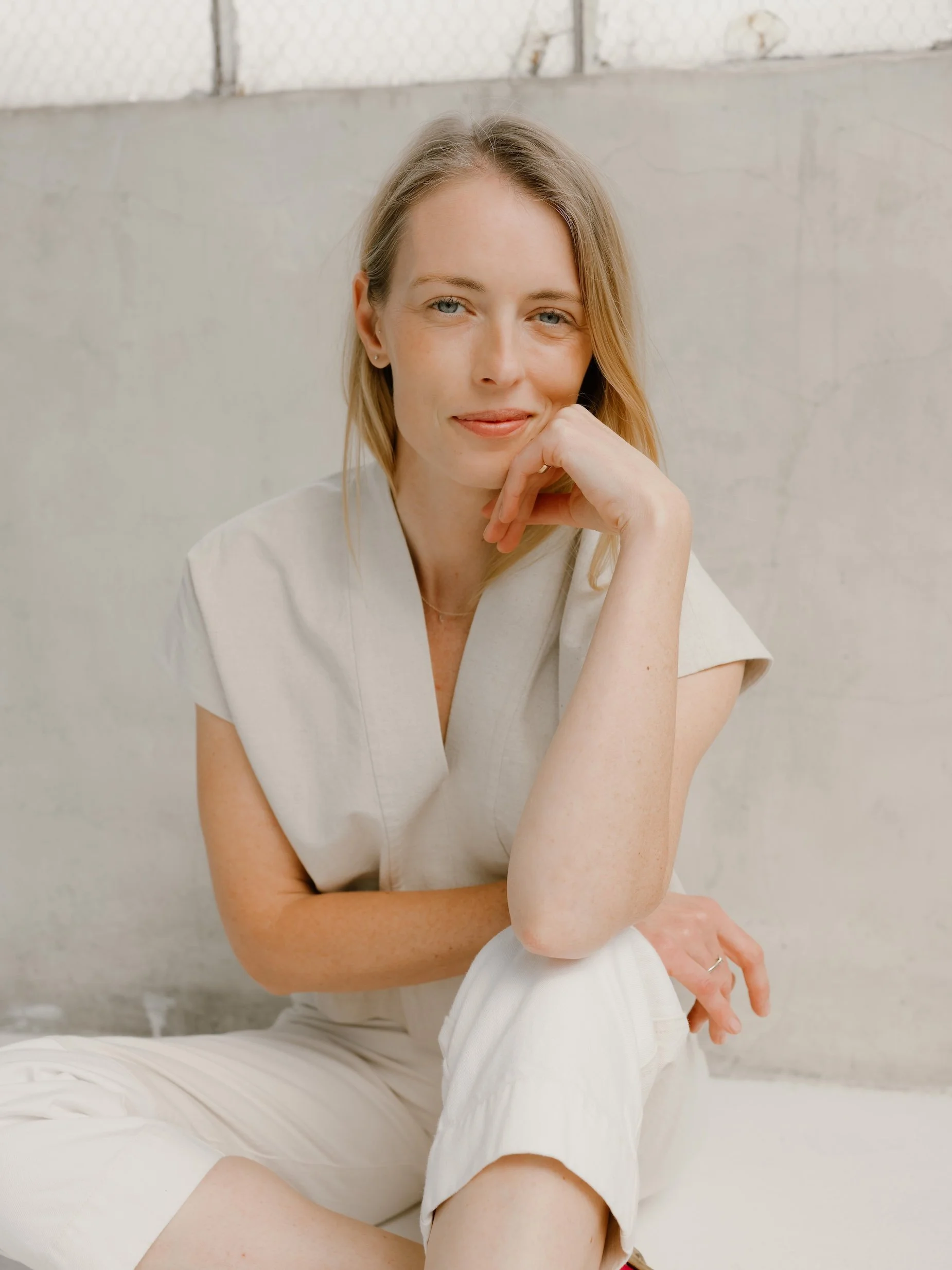 A woman with light skin and blonde hair sits on a bed, resting her chin on her hand, in front of a concrete wall.