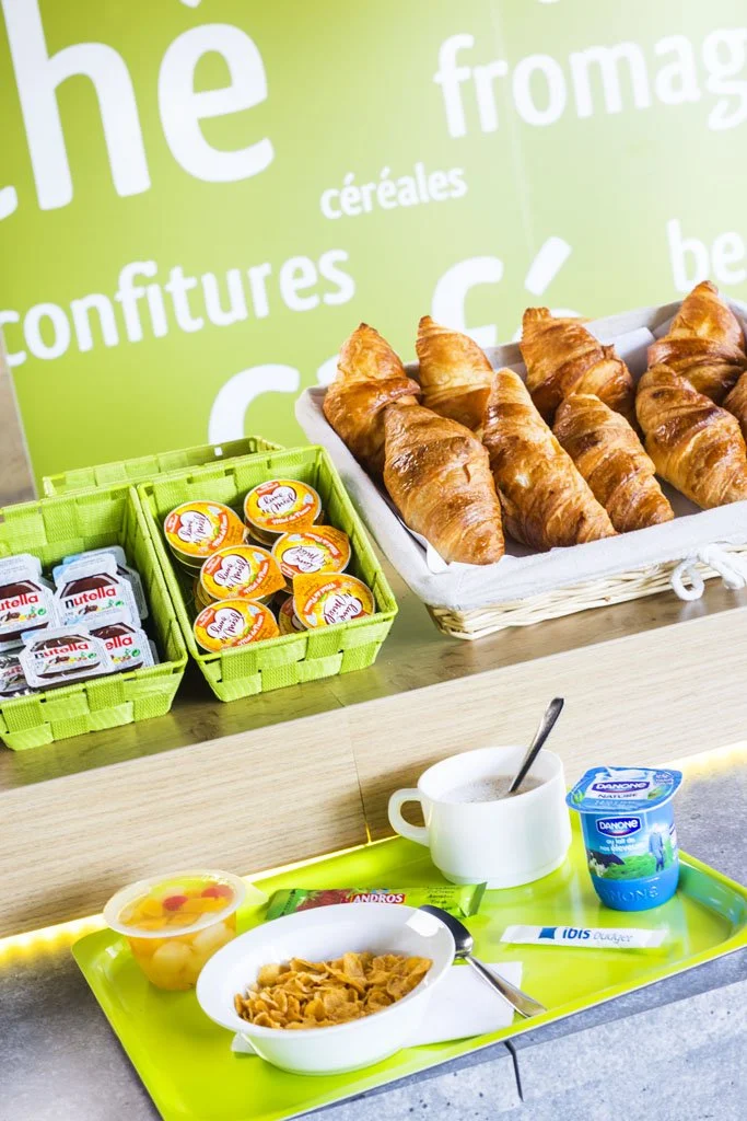 Desayuno con cruasanes, mermeladas, Nutella, yogur, cereales, una taza de café o té y un bol de fruta. Paredes verdes al fondo con palabras en francés como «fromage», «céréales» y «confitures».