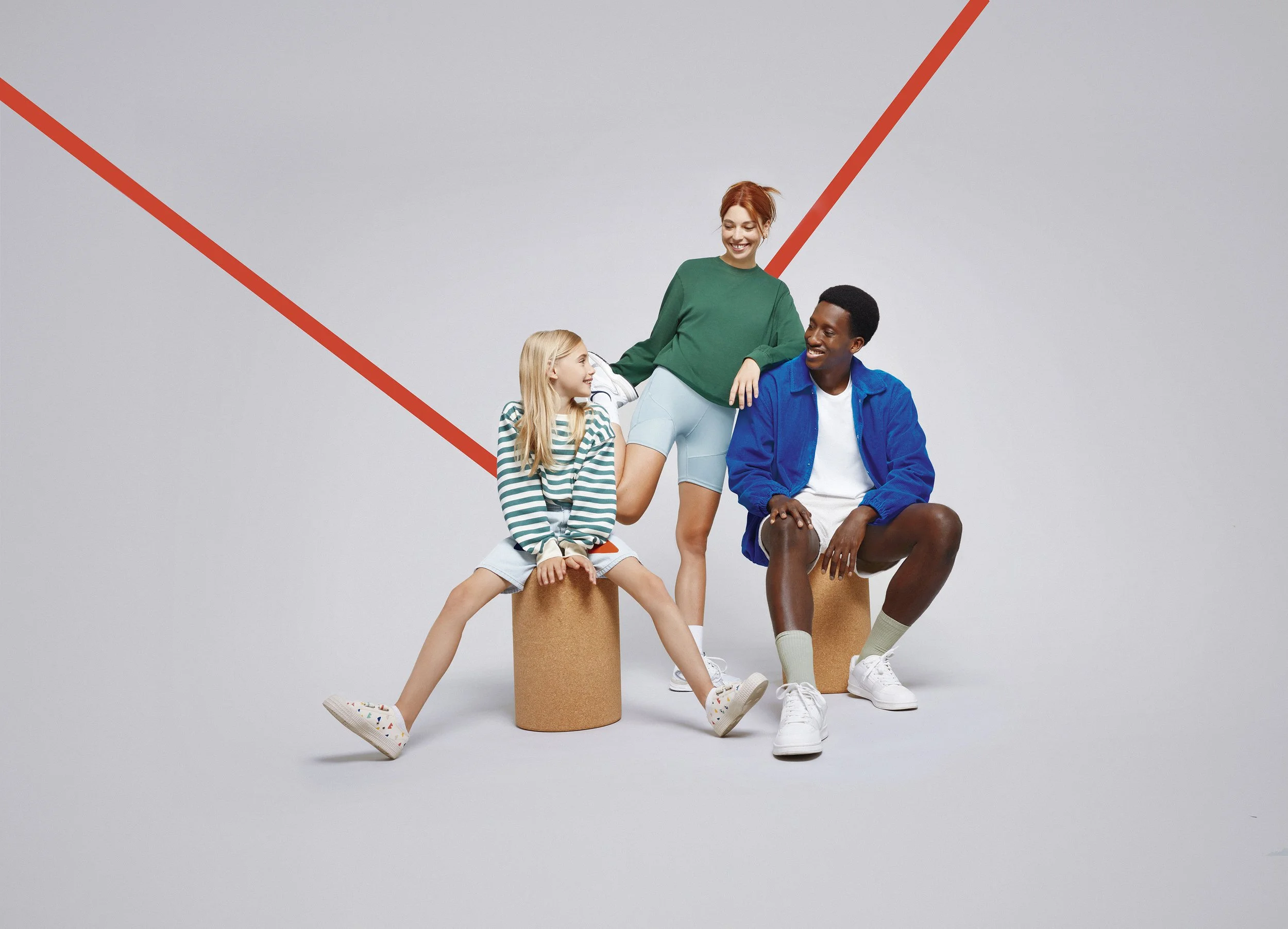 Trois personnes, deux femmes et un homme, assises et debout sur des blocs en carton, souriant et discutant ensemble. Fond blanc avec deux lignes rouges diagonales.