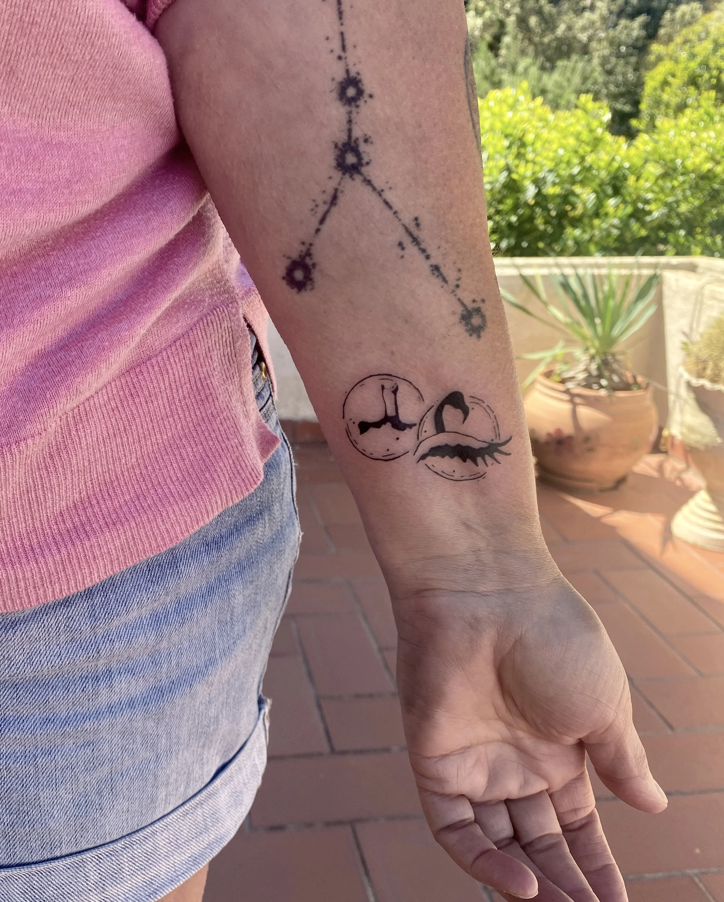 Tatouage d'une constellation avec cinq étoiles sur le bras d'une personne, arrière-plan de plantes en pot et ciel ensoleillé.
