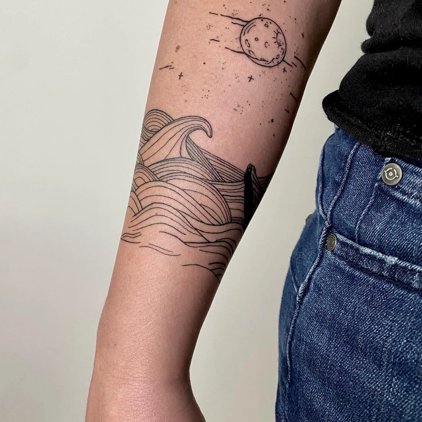 Tatouage en ligne représentant des vagues sur le bras d'une personne, sous un dessin de la lune avec des étoiles.