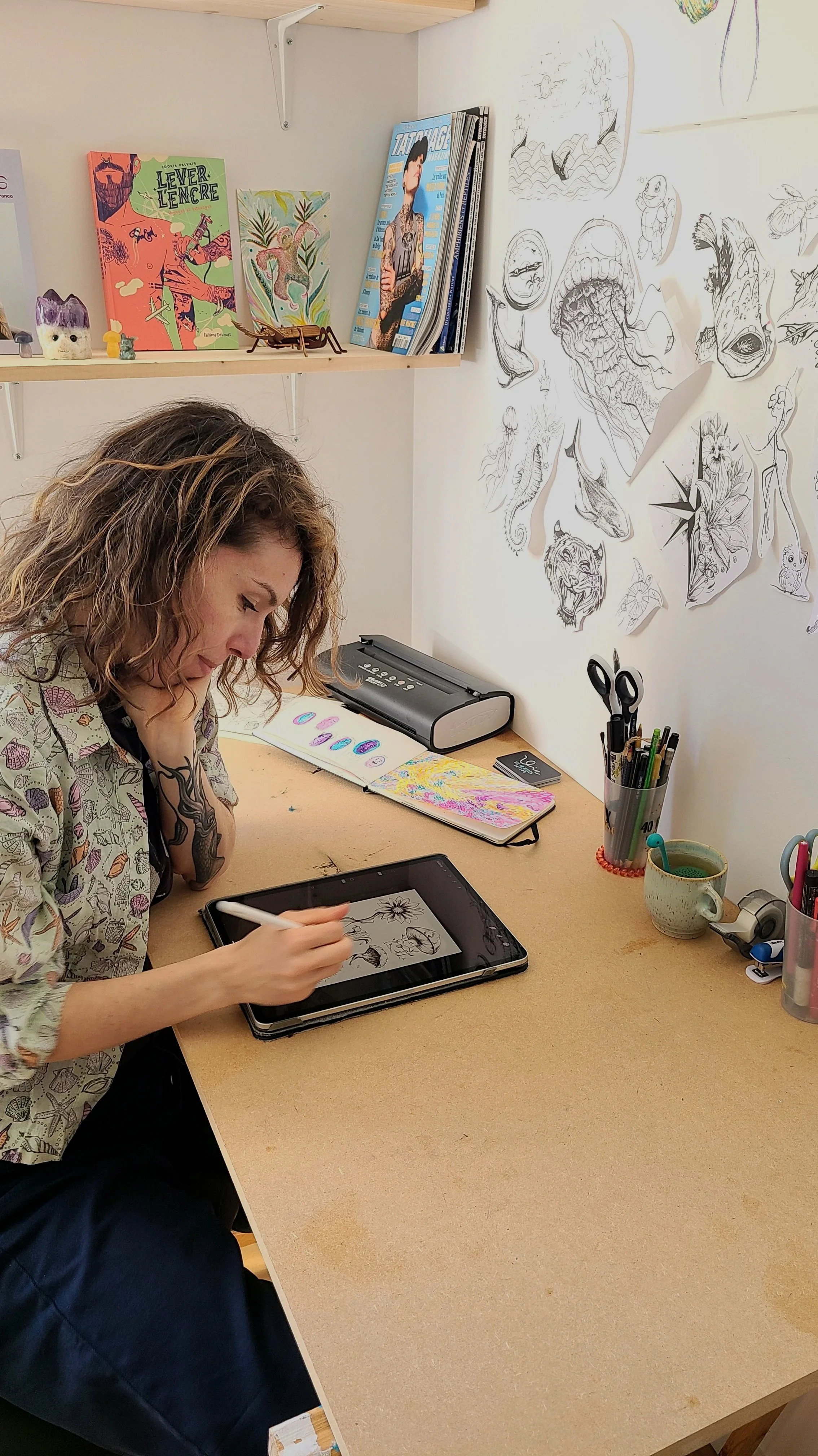 Une femme assise à un bureau en train de dessiner ou de peindre sur une tablette graphique, entourée de fournitures d'art et de tatouages en cours sur ses bras.