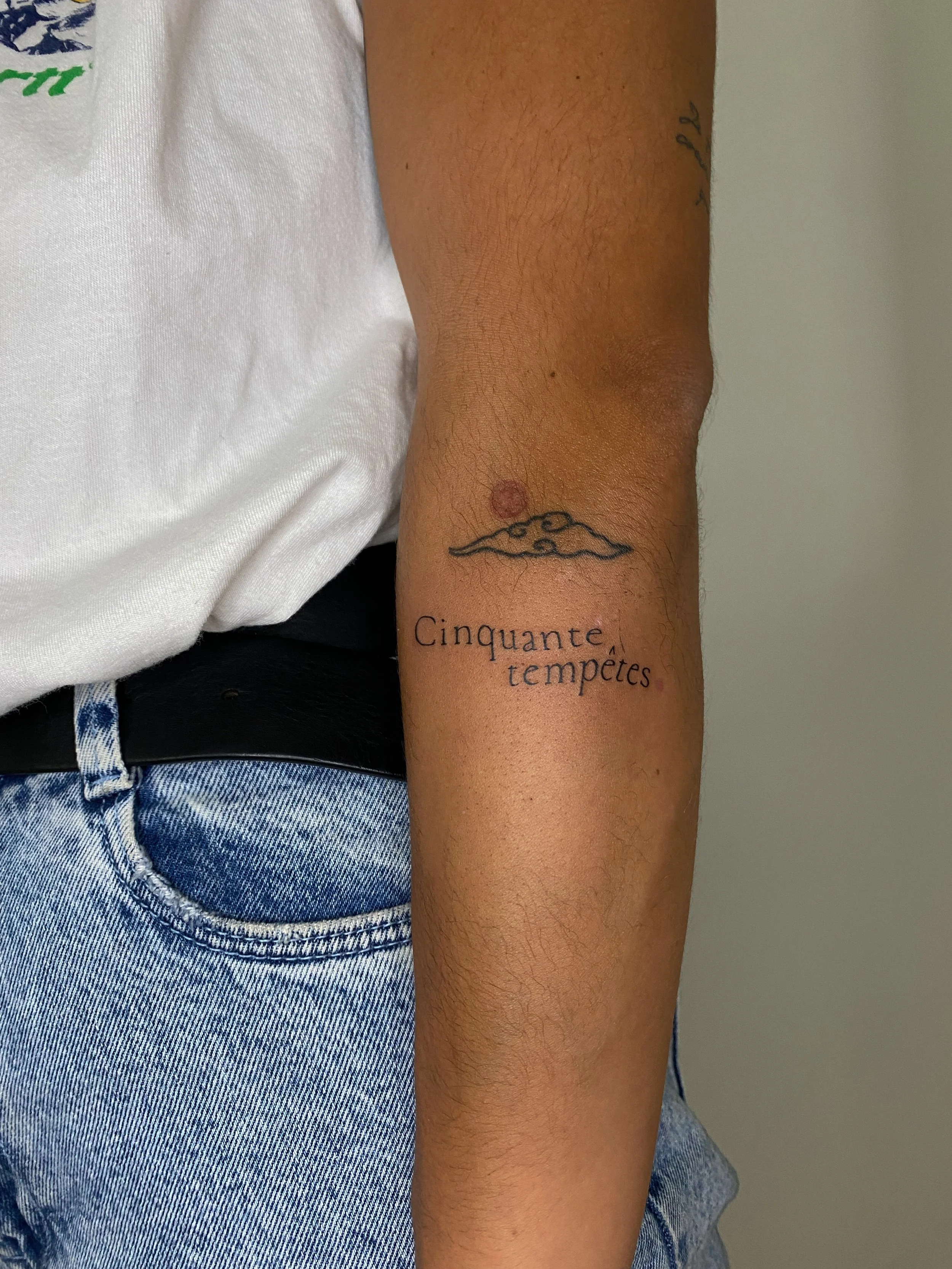 Tatouage d'une montagne avec un soleil derrière sur le bras gauche d'une personne, portant un t-shirt blanc et un jean bleu.