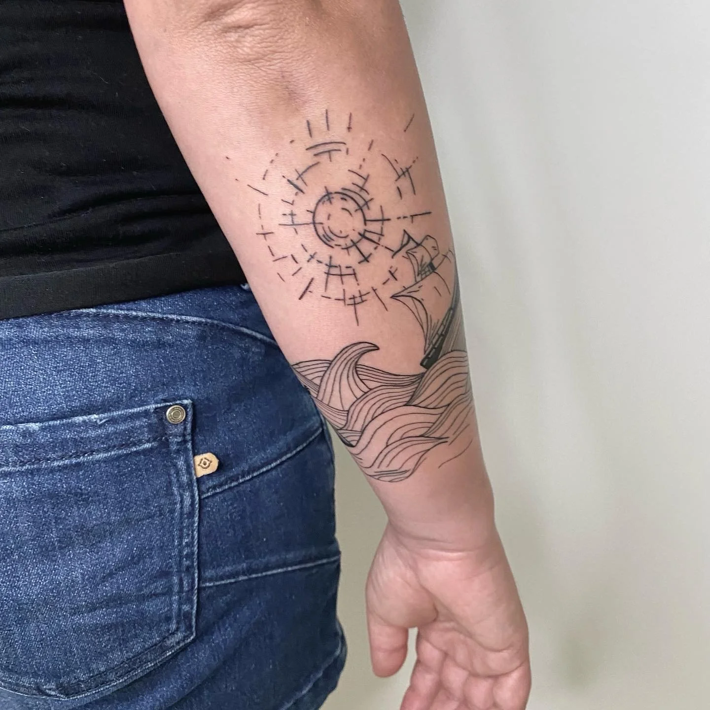 Tatouage d'un bateau avec des vagues, un soleil et des lignes stylisées sur le bras d'une personne.