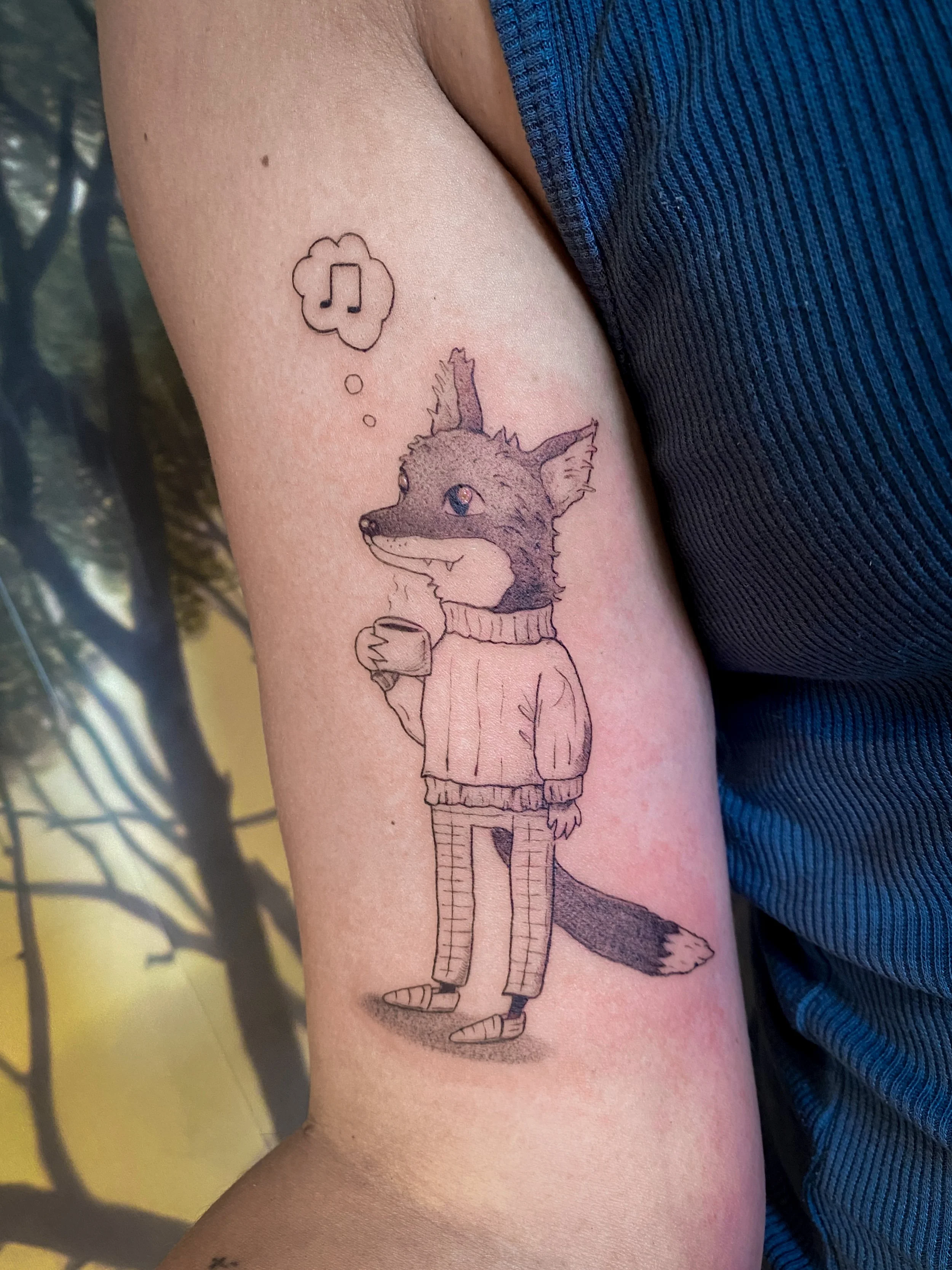 Tatouage dessiné d'un renard humain portant un pull, un pantalon rayé et des chaussettes, tenant une tasse, avec une bulle de pensée contenant une note de musique.