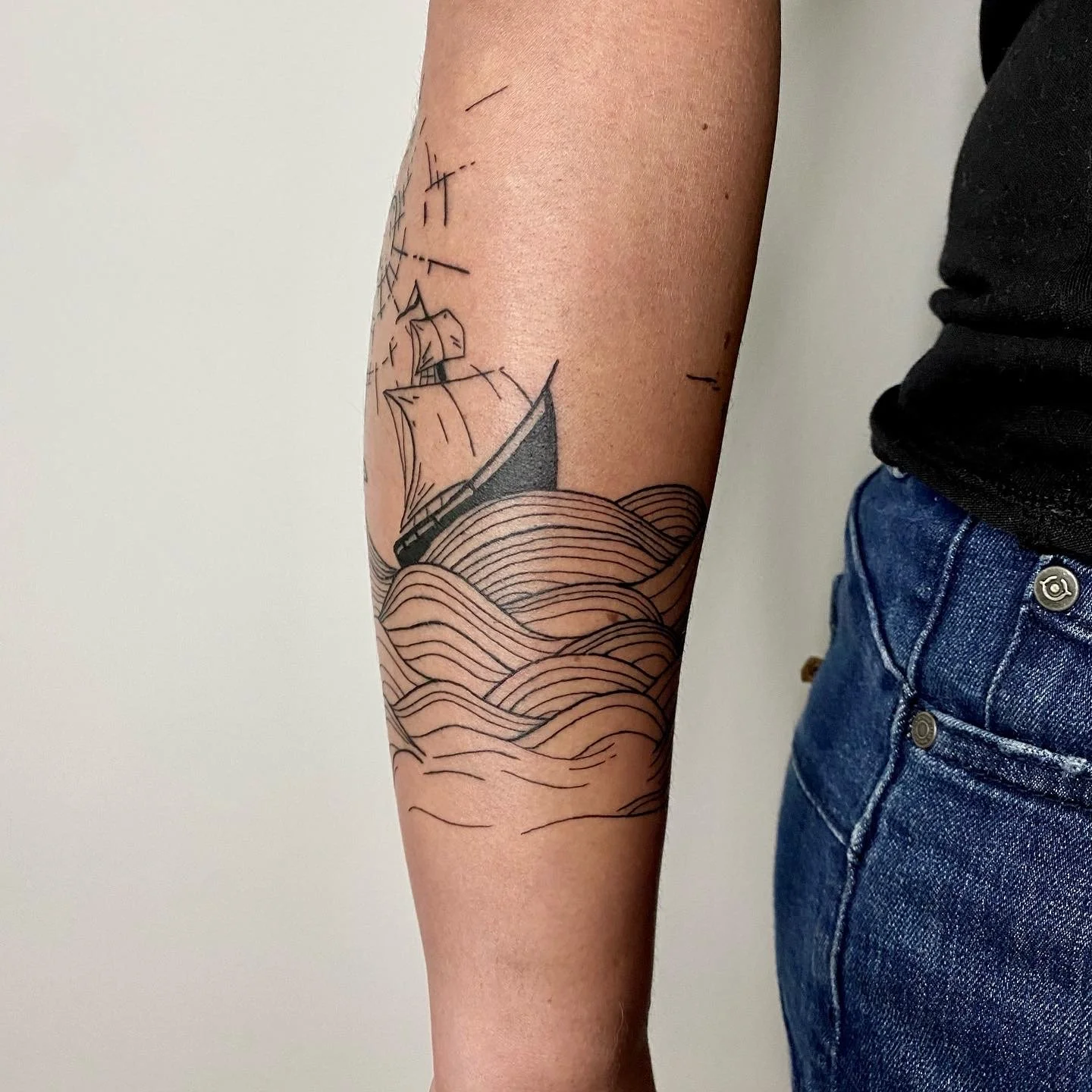 Tatouage d'un bateau naviguant sur des vagues sur le bras d'une personne, avec une tenue décontractée et un fond neutre.
