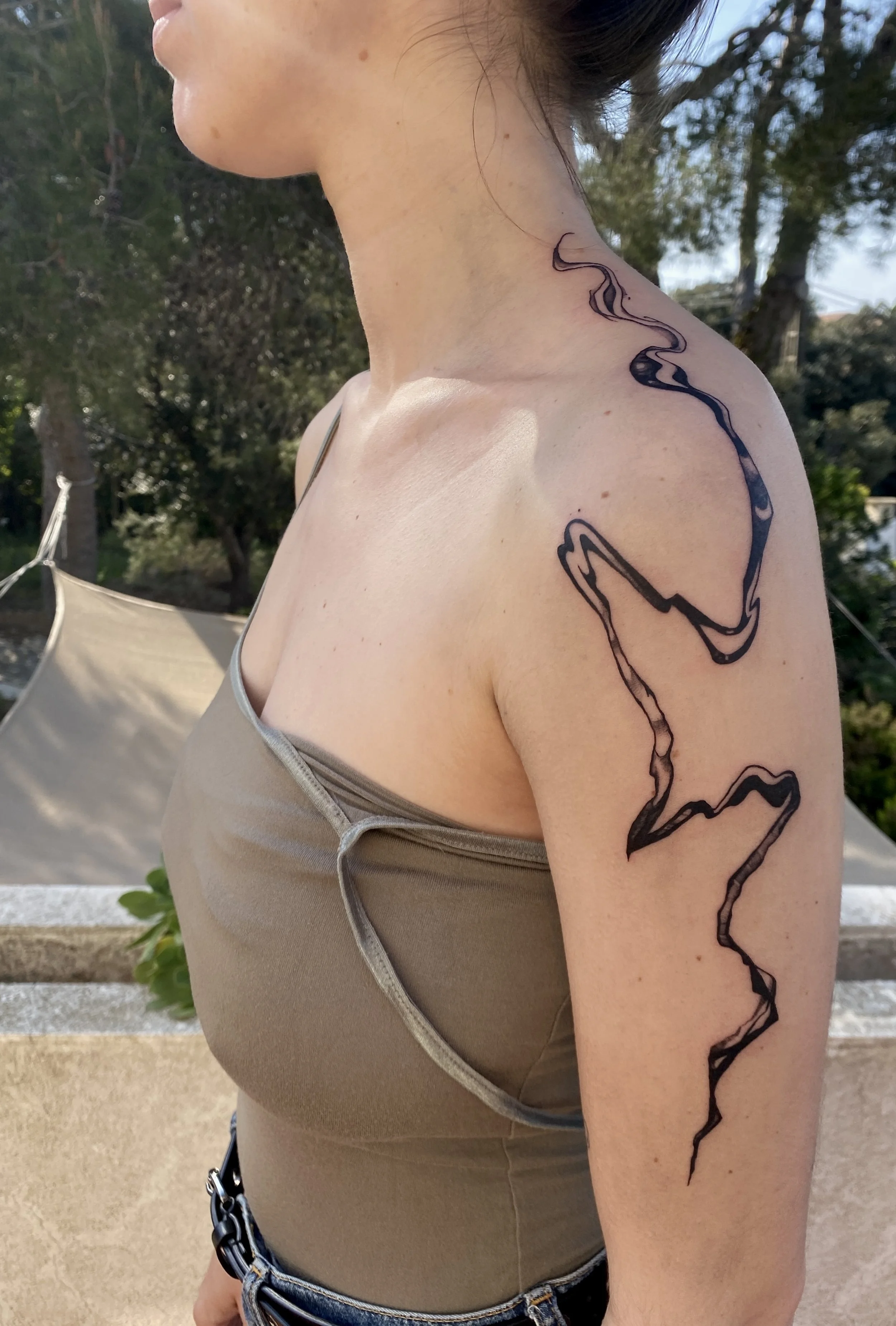 Une femme porte une robe beige avec un décolleté en V et une chaîne noire. Elle a un tatouage noir en forme de ligne sinueuse sur l'épaule et le bras. Le décor extérieur comporte des arbres et un parasol beige en arrière-plan.