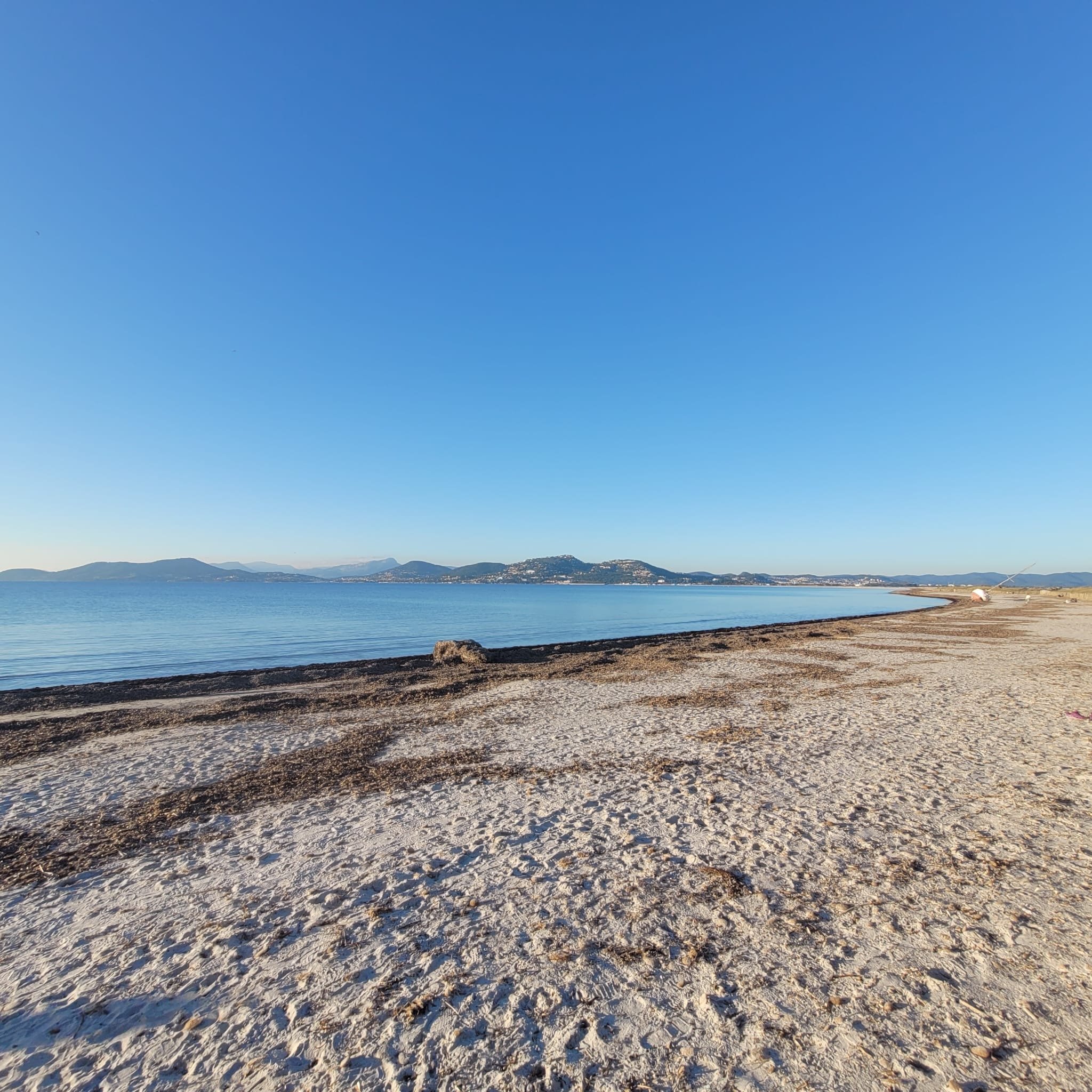 Plage de sable avec des rochers, vue sur un lac ou une mer, montagnes en arrière-plan, ciel clair et ensoleillé.