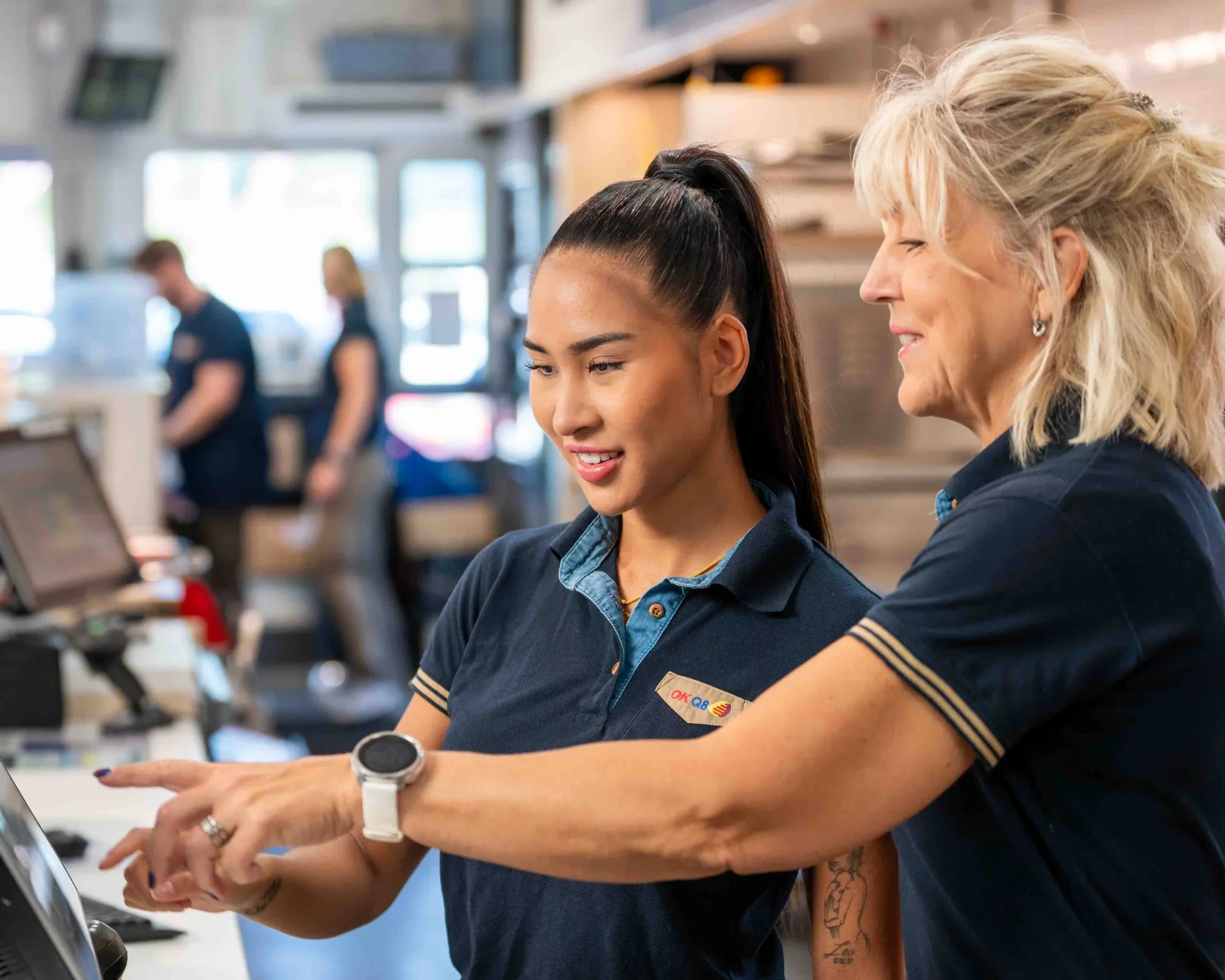 At a checkout counter, a young female cashier with a name tag and a senior female customer with blonde hair and earrings are smiling and looking at a digital screen, engaging in a friendly conversation. In the background, other employees are working.