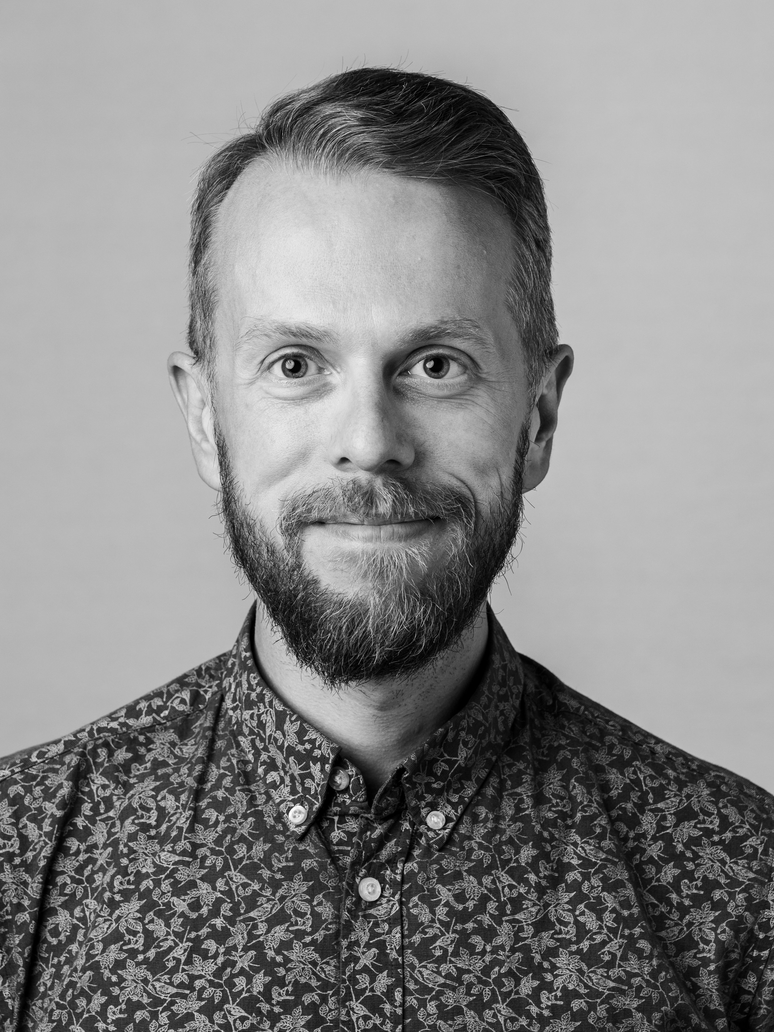 Black and white portrait of a man with short, slicked-back hair, a beard, and wearing a patterned shirt.
