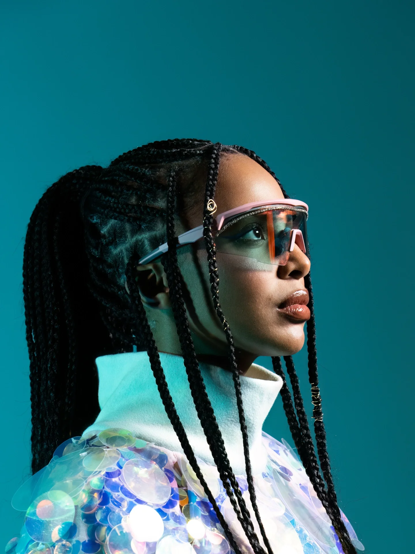 A woman with braided hair and reflective goggles facing right, wearing a colorful, sequined garment against a teal background.