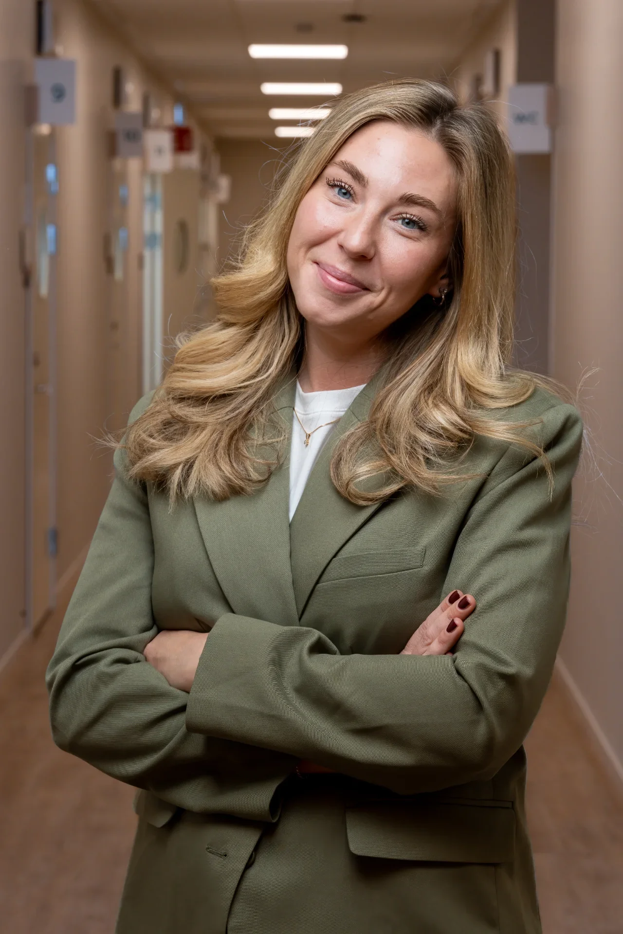 A young woman with long blonde hair and blue eyes standing in a hallway, smiling with her arms crossed, wearing a green blazer and white top.