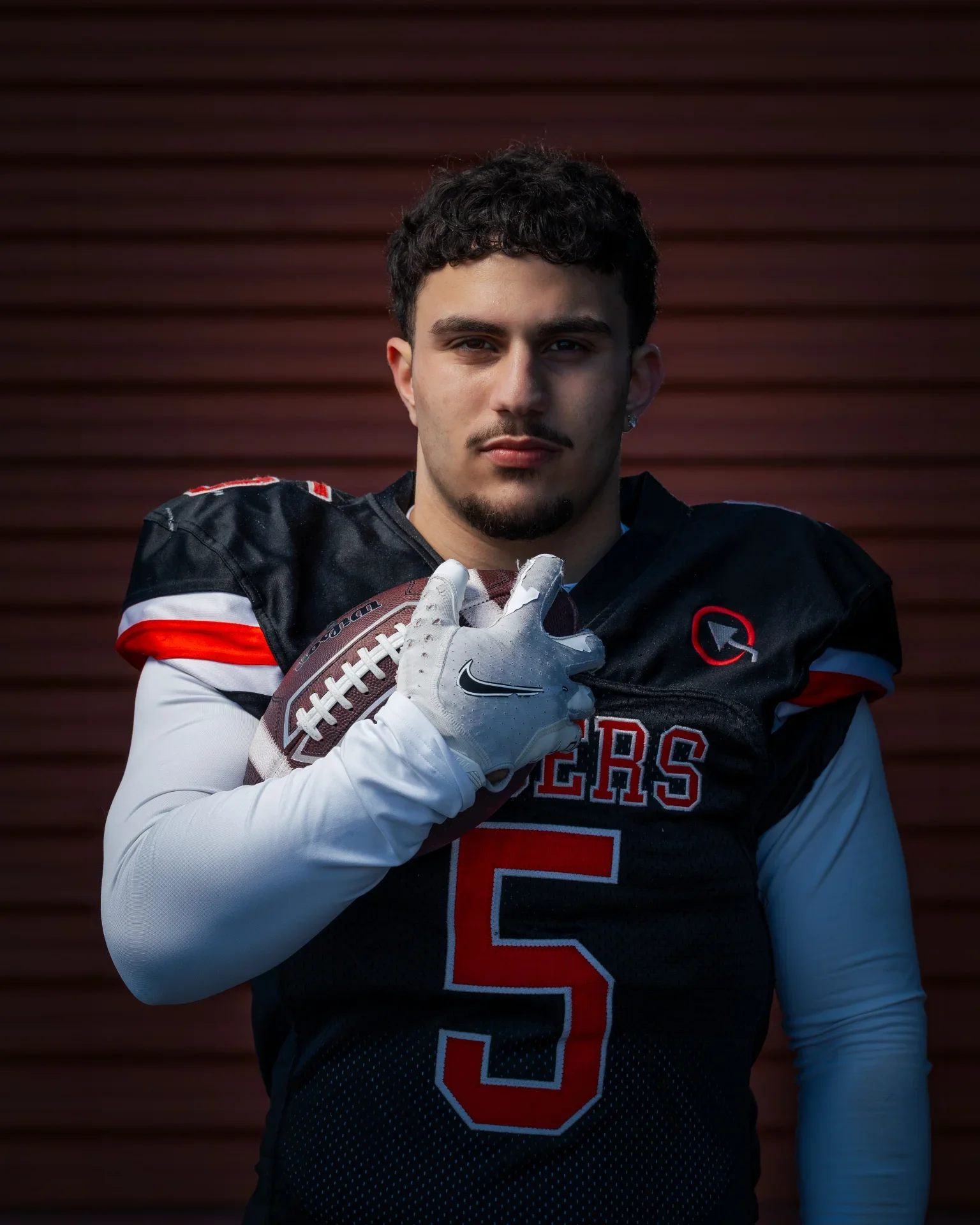Young football player with dark hair and a goatee holding a football against a brown horizontal slatted background, wearing a black and white football jersey with red accents and a red number 3.