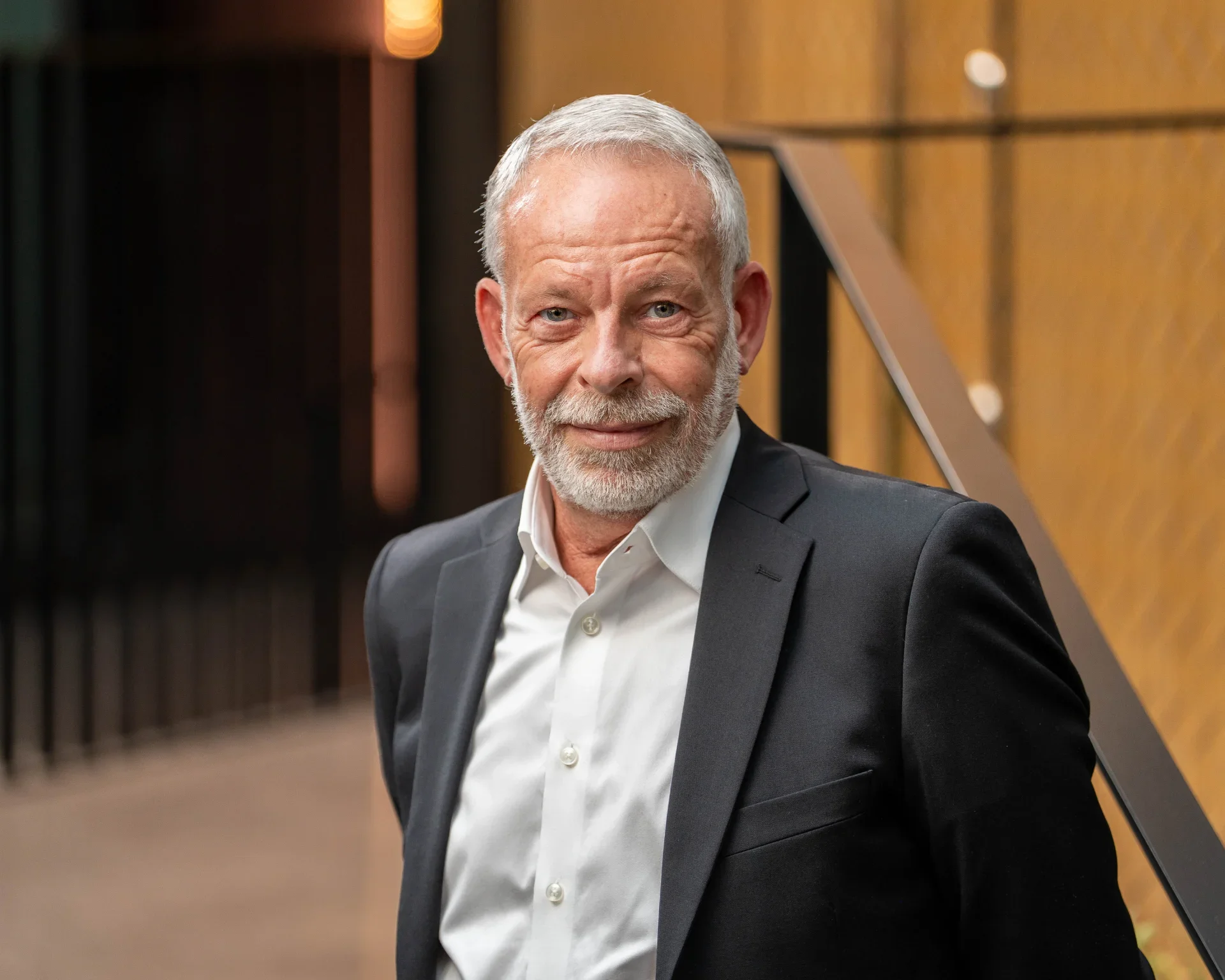 A smiling older man with a beard, dressed in a dark suit and white shirt, standing indoors near a staircase with wood paneling.