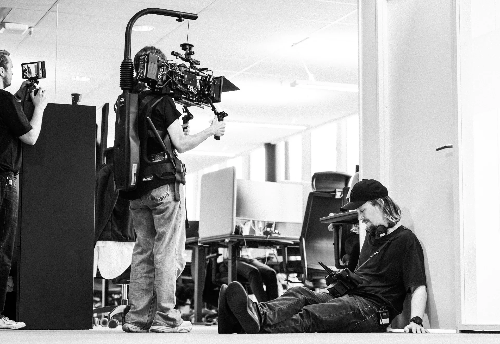 Filmmaking crew on set, one person filming with a camera rig, person sitting on floor with a device, and another person organizing equipment.